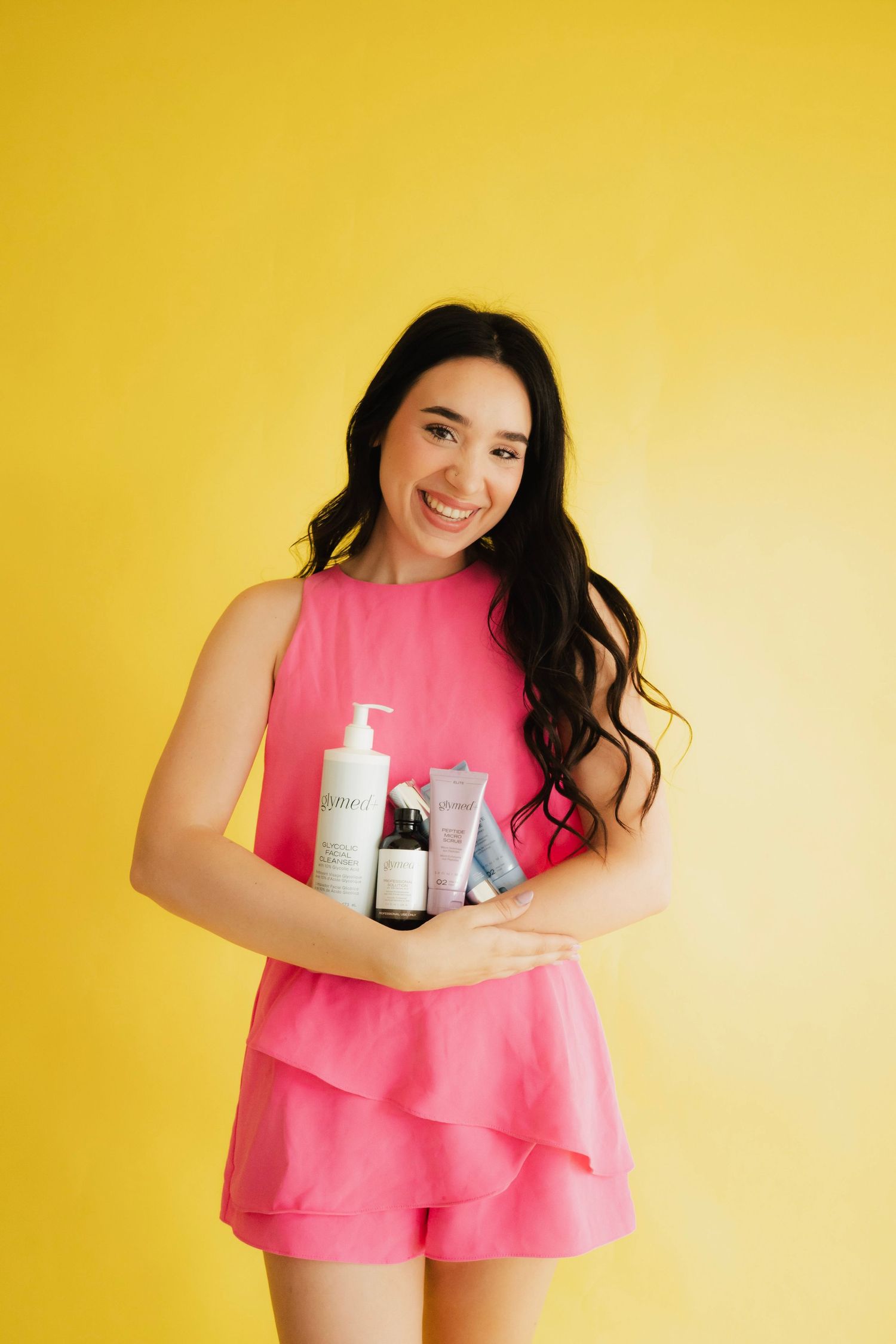 Smiling woman in pink romper holding skincare products against a bright yellow background.