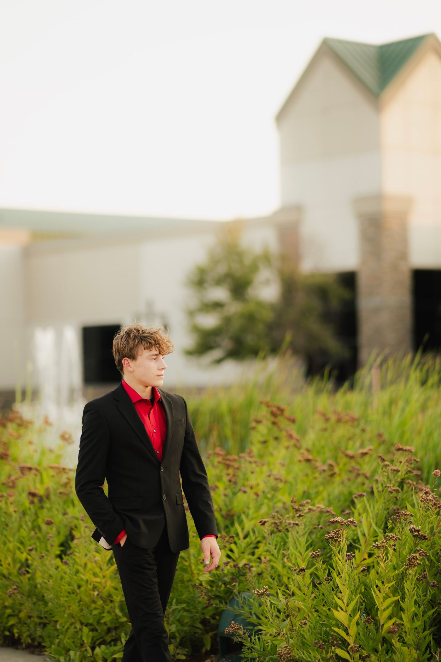 Young man in black suit and red shirt walking through lush green garden outside elegant building.