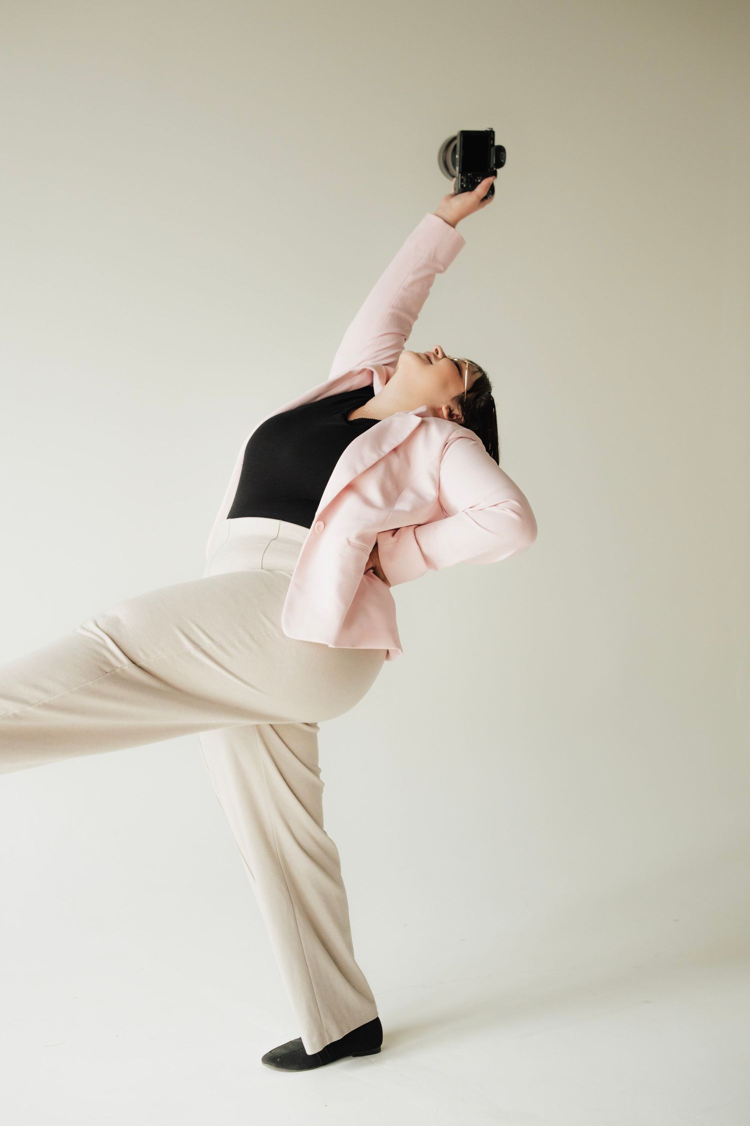 Woman in pink blazer dramatically poses with camera raised overhead against white background.