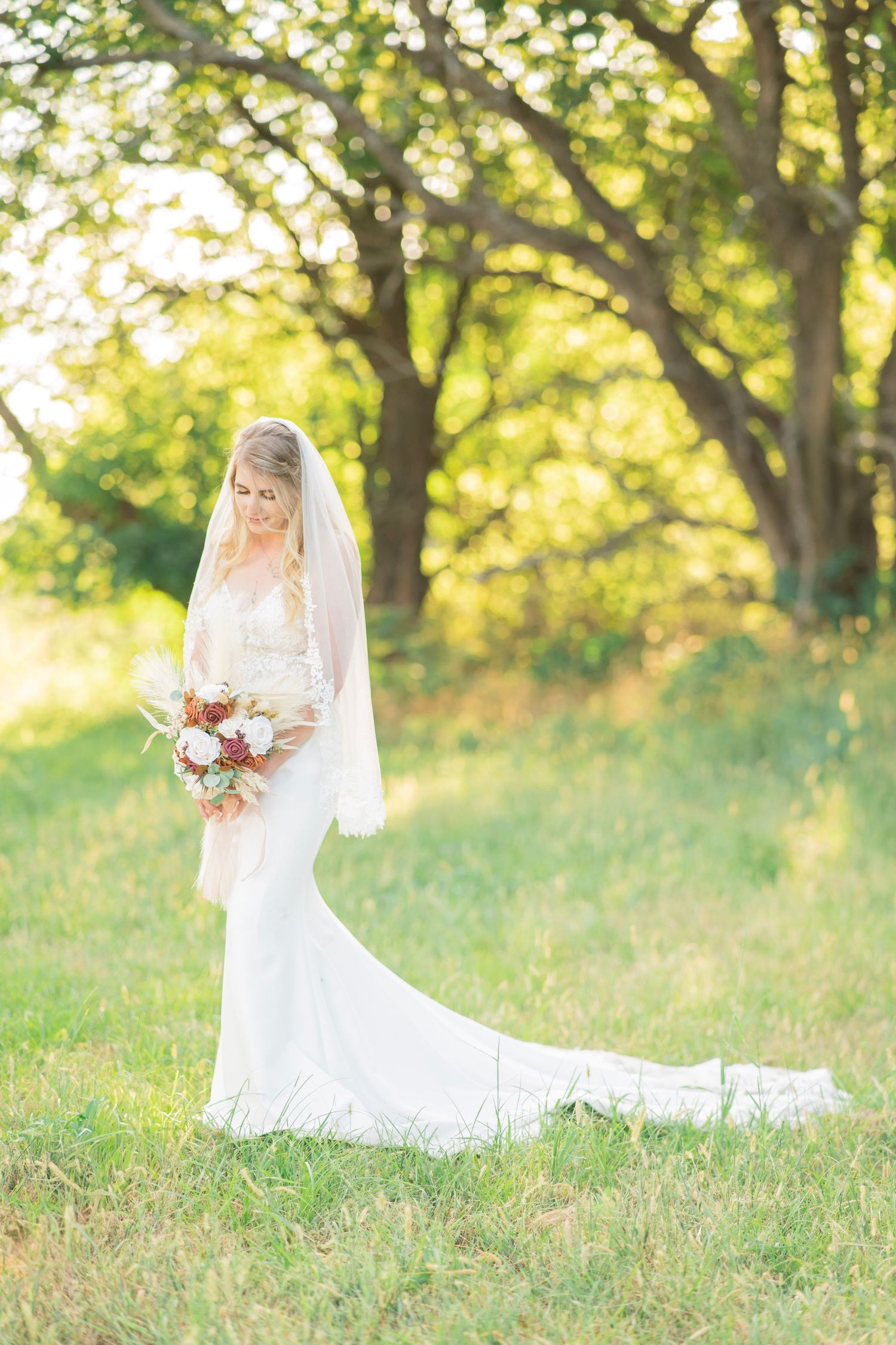 Elegant bride in lace gown and long veil holding floral bouquet in sunlit green outdoor field.