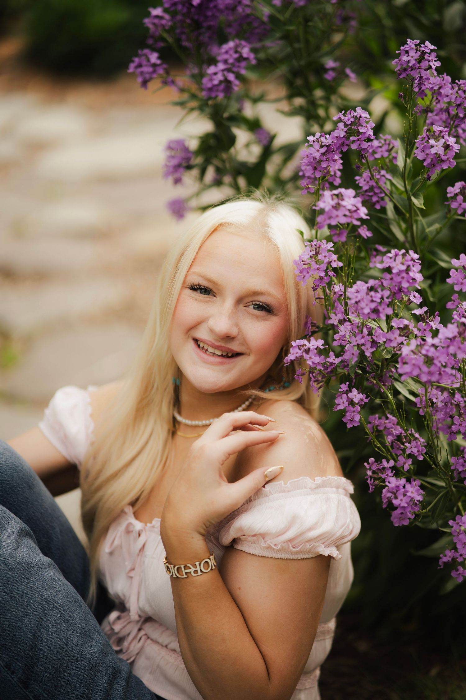 Smiling blonde girl in pink off-shoulder top seated beside vibrant purple flowers in a garden.