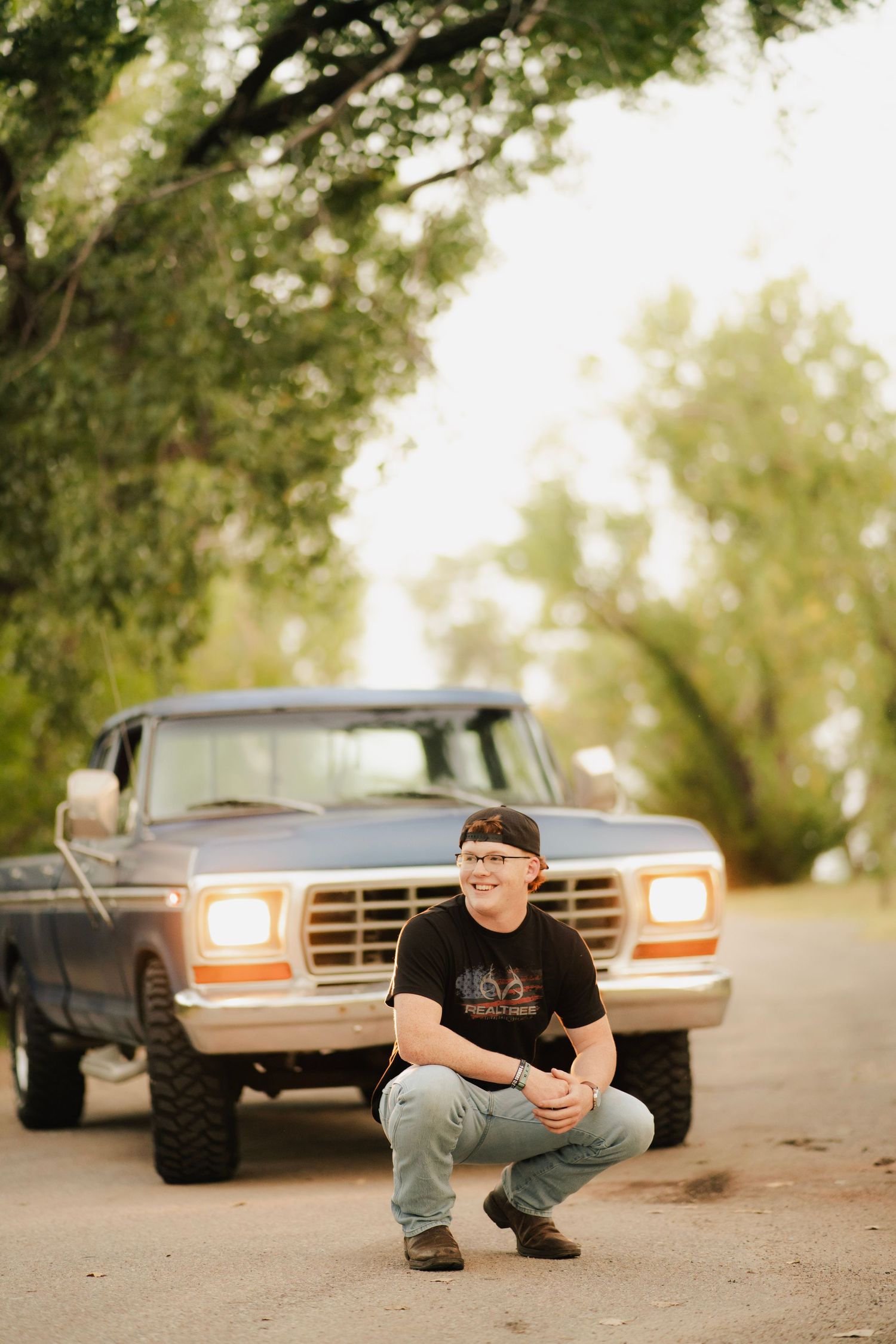 Young man crouching in front of a classic Ford pickup truck on a tree-lined dirt road.