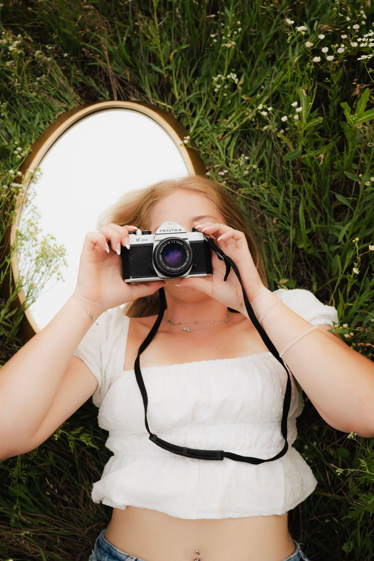 Blonde girl lying in wildflower field holding vintage film camera up to her face.