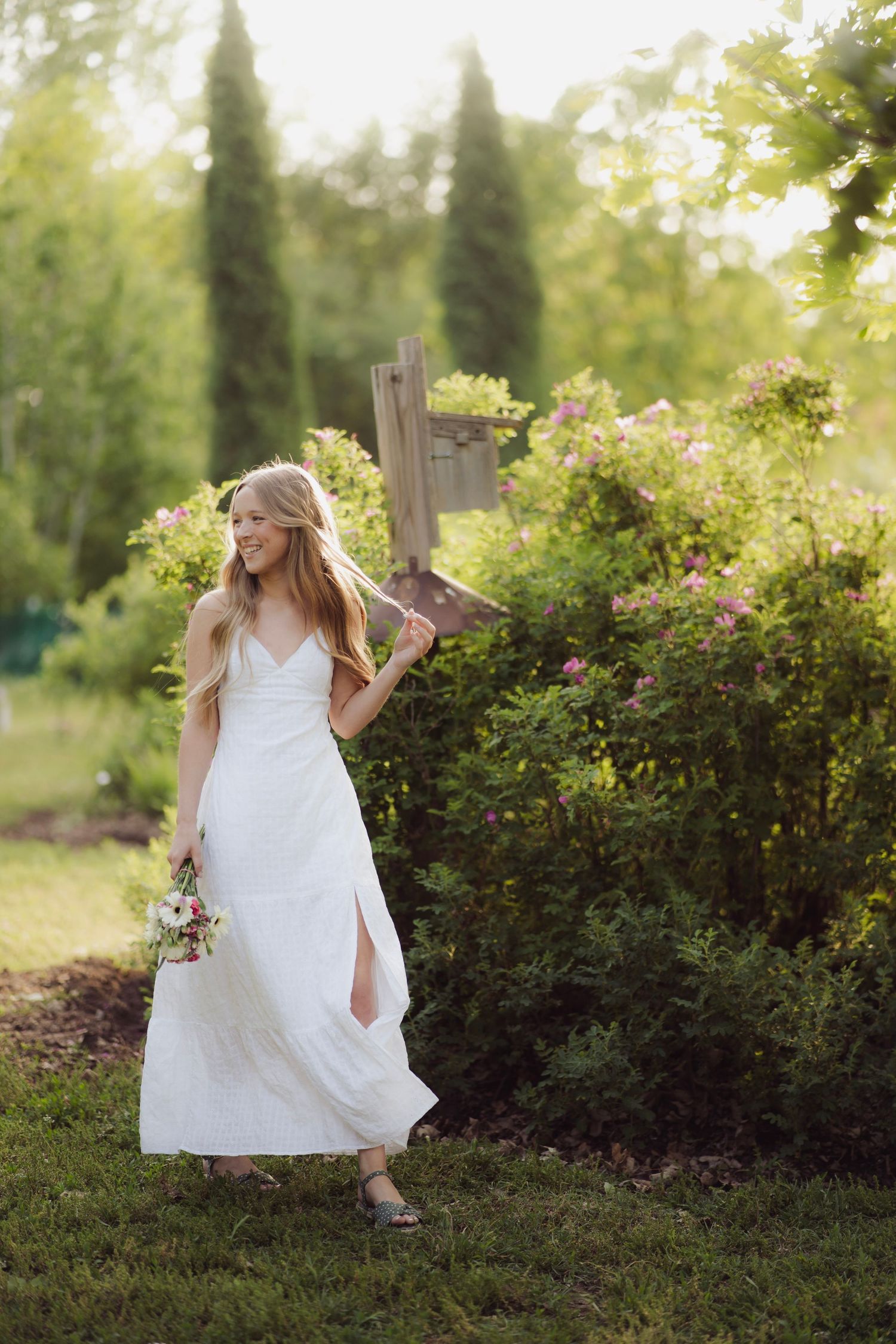 Blonde woman in white maxi dress holding bouquet and wooden sign in sunny garden.