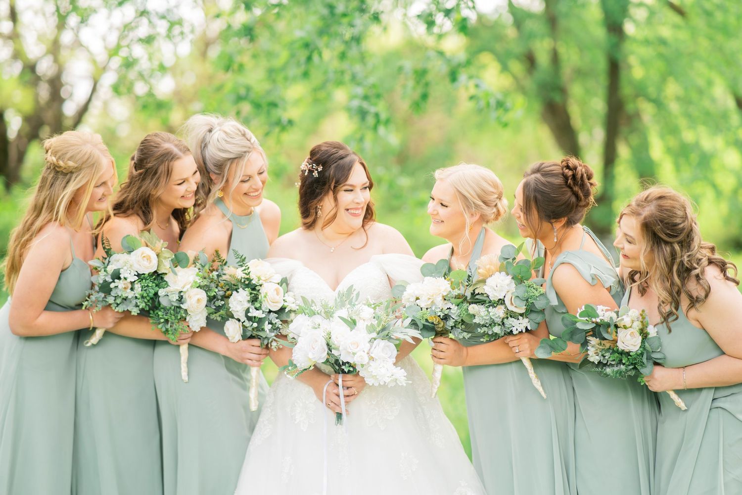 Bride and six bridesmaids in sage green dresses laugh together outdoors holding white floral bouquets.