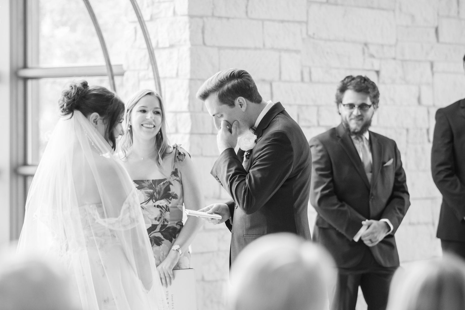 Emotional groom wipes tears during wedding ceremony while officiant and bride smile in black and white photo.