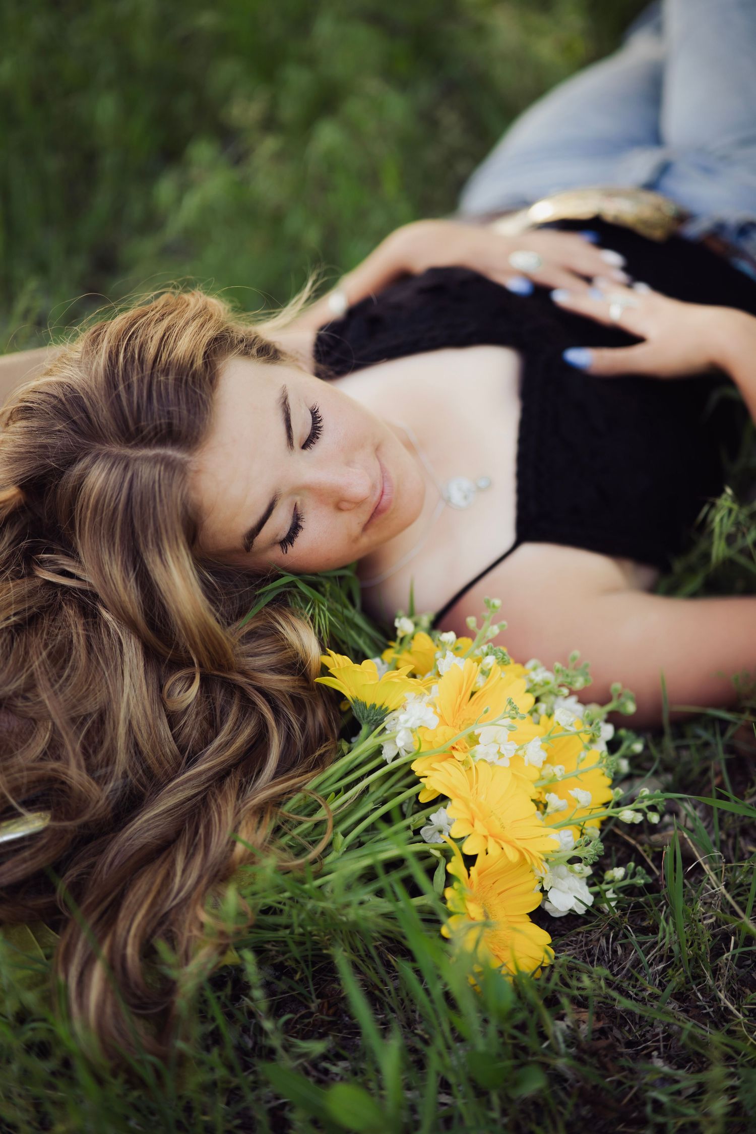 Young woman with wavy hair lies in green grass beside yellow and white wildflower bouquet outdoors.