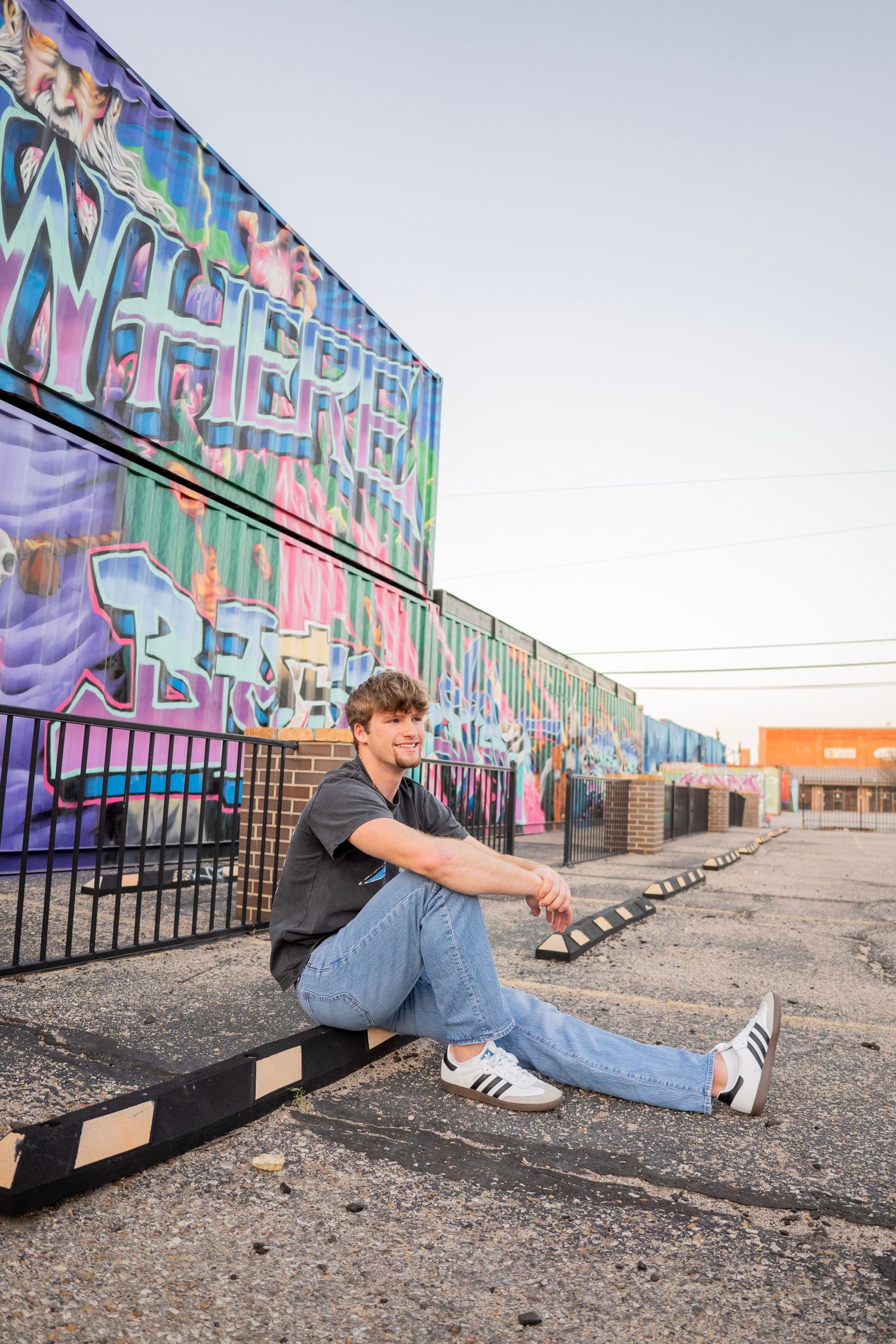 Young man in jeans and white sneakers sitting on a parking curb in front of a colorful graffiti mural wall.