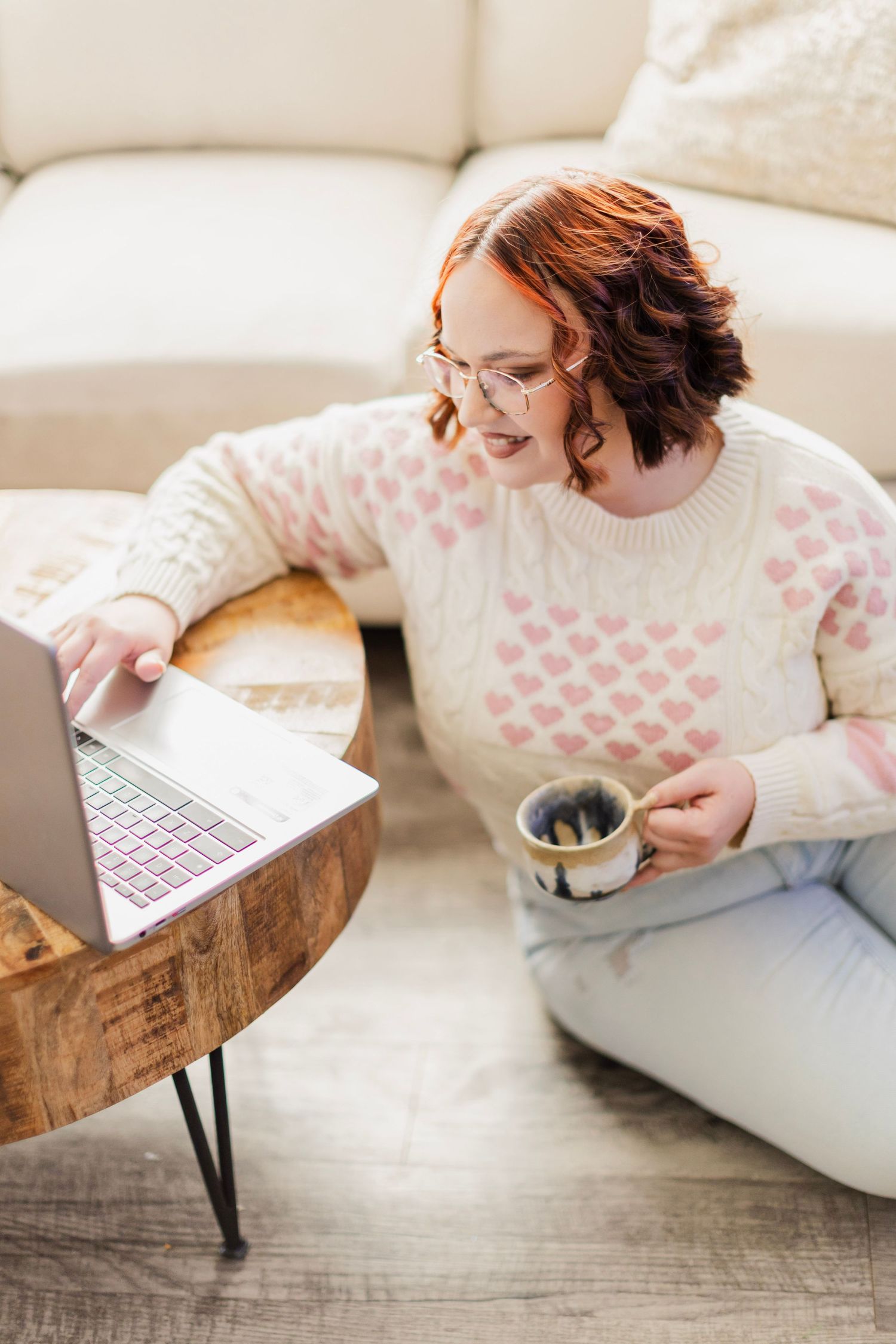 A woman in a heart-print sweater sits on the floor working on a laptop while holding a coffee mug.
