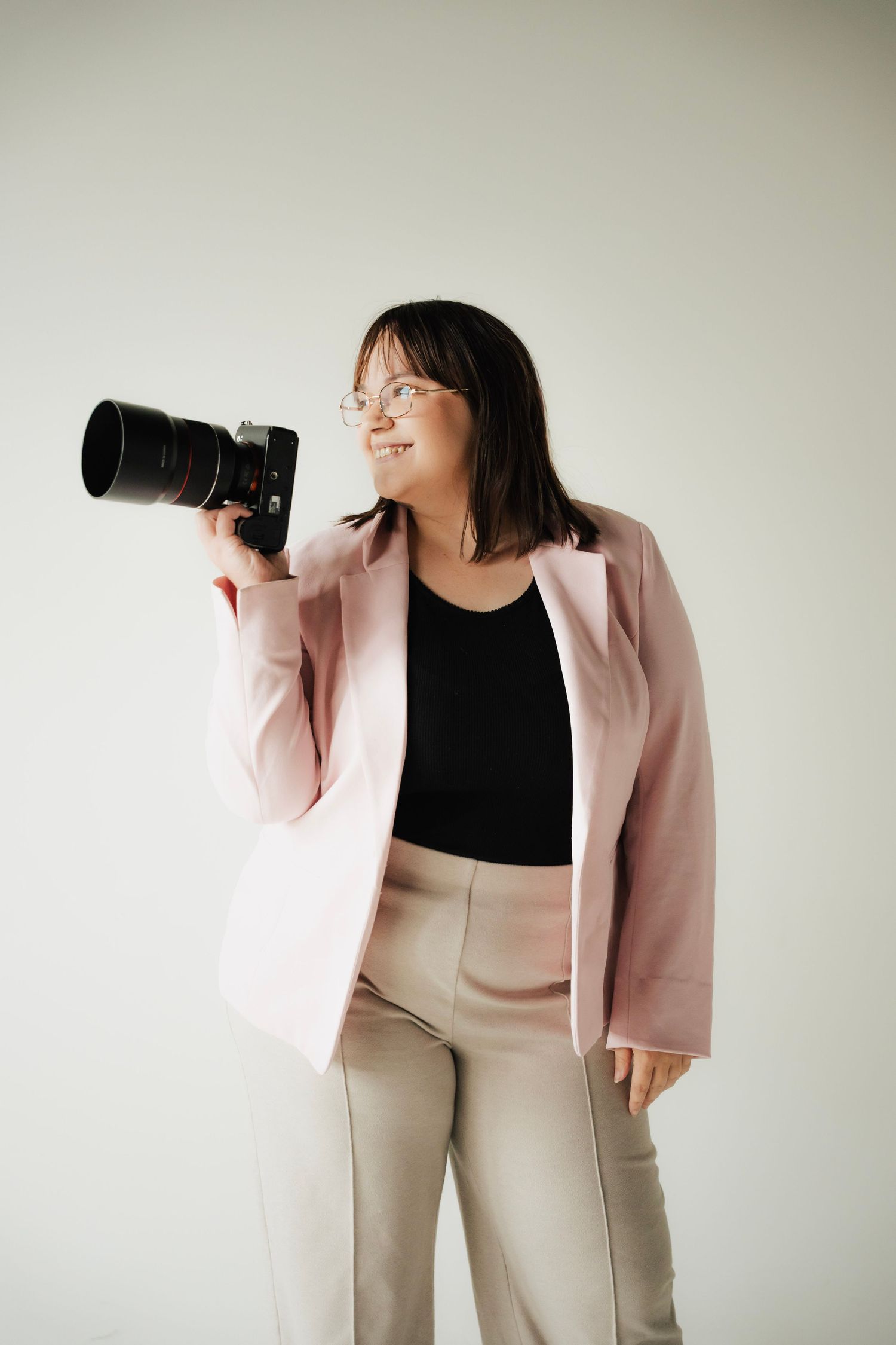 Professional female photographer in pink blazer holding DSLR camera while looking away confidently.