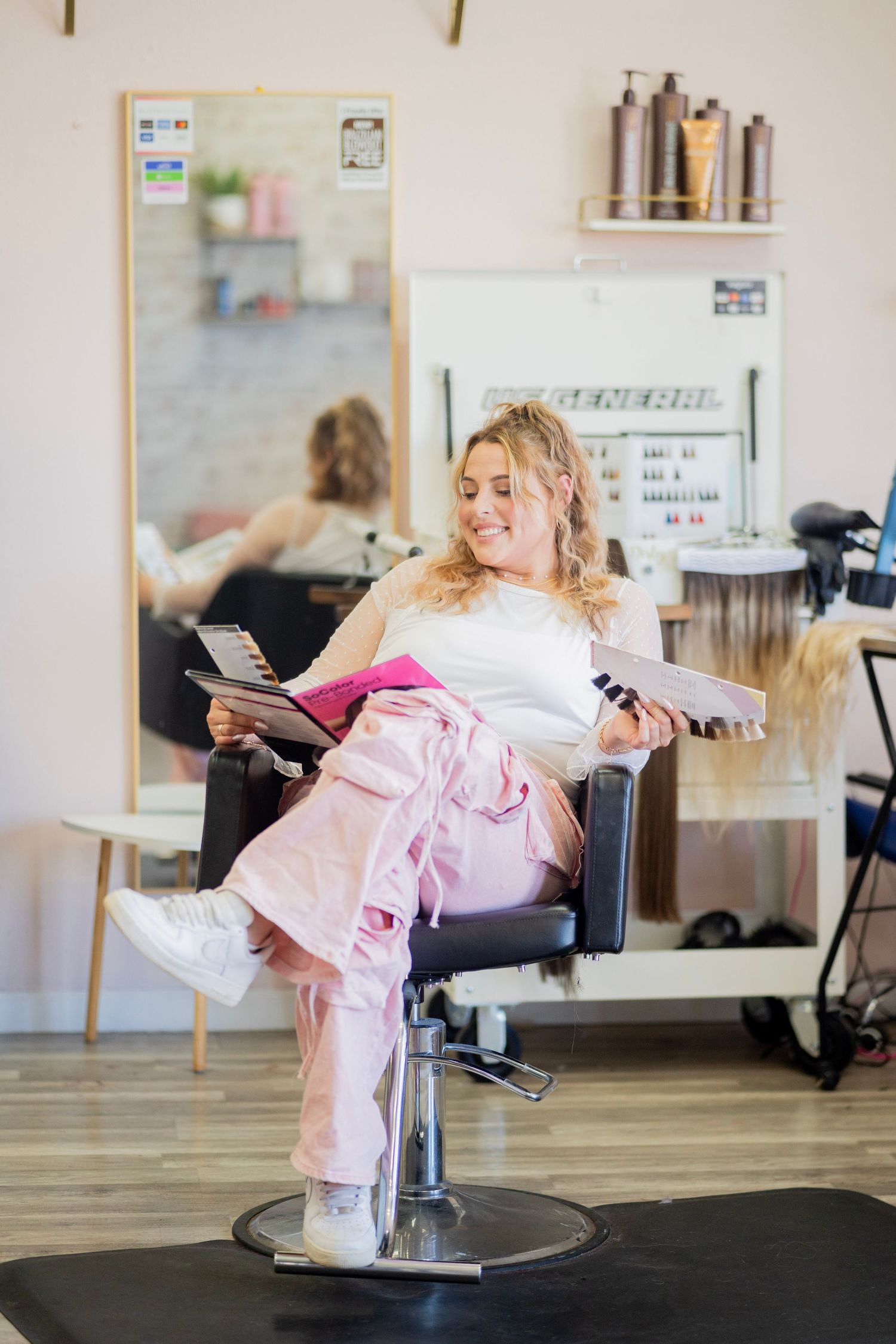 Smiling blonde woman sits in salon chair holding color swatches in a bright hair salon.