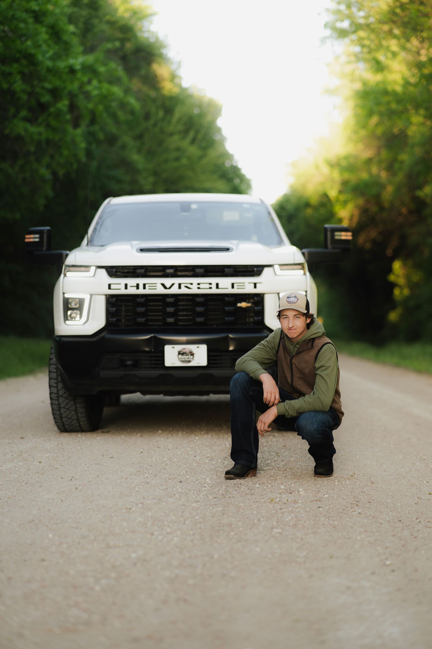 Young man crouching in front of a white Chevrolet pickup truck on a rural gravel road surrounded by green trees.