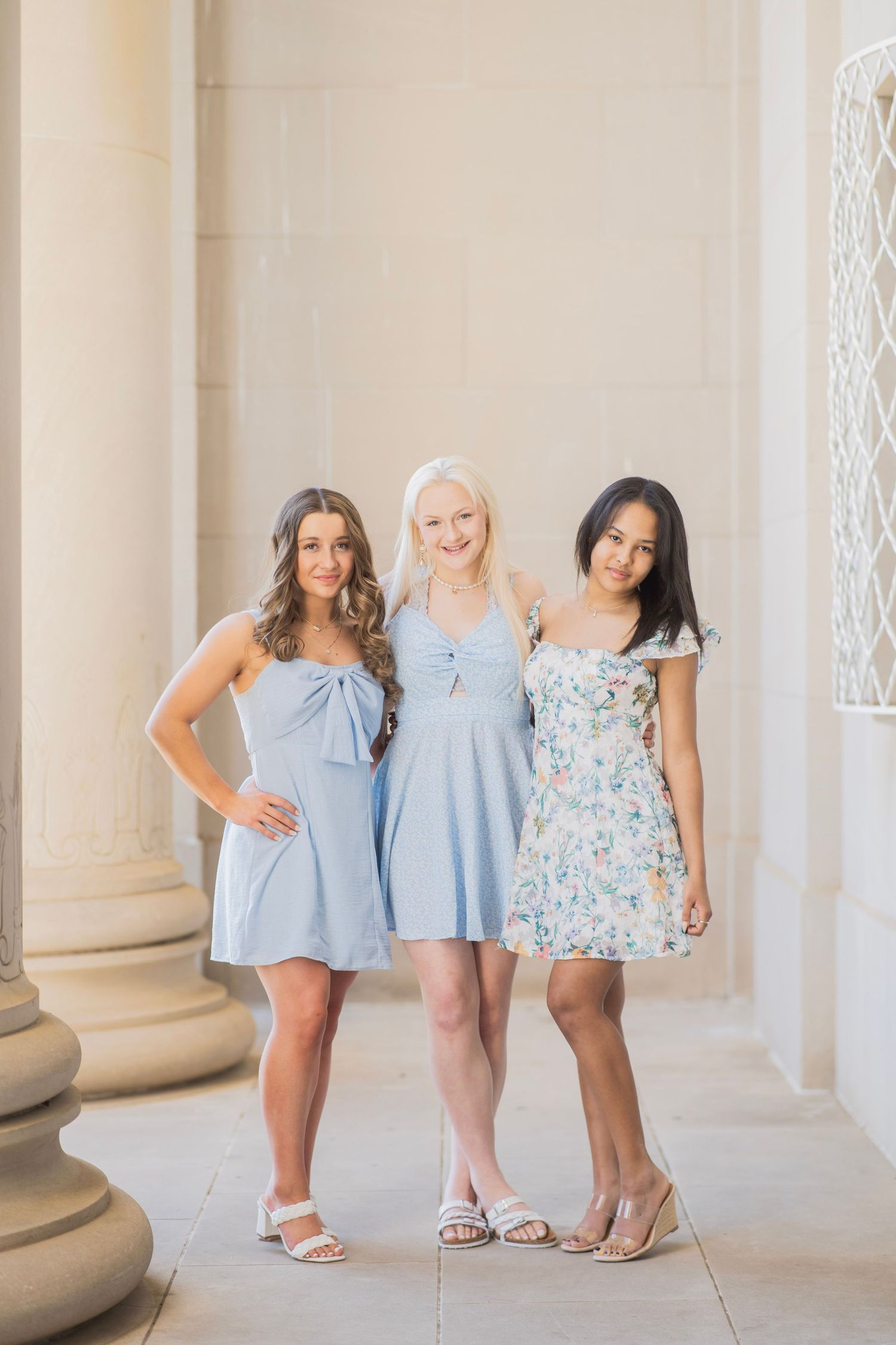 Three teenage girls in light blue and floral dresses pose together between stone columns outdoors.
