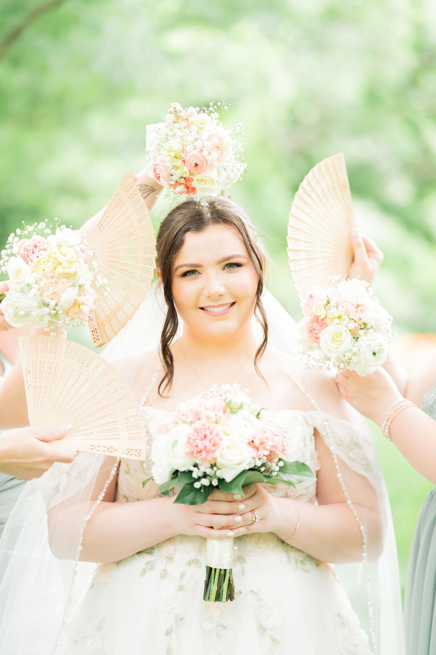 Smiling bride holding pink and white bouquet surrounded by bridesmaids with decorative fans at outdoor wedding.