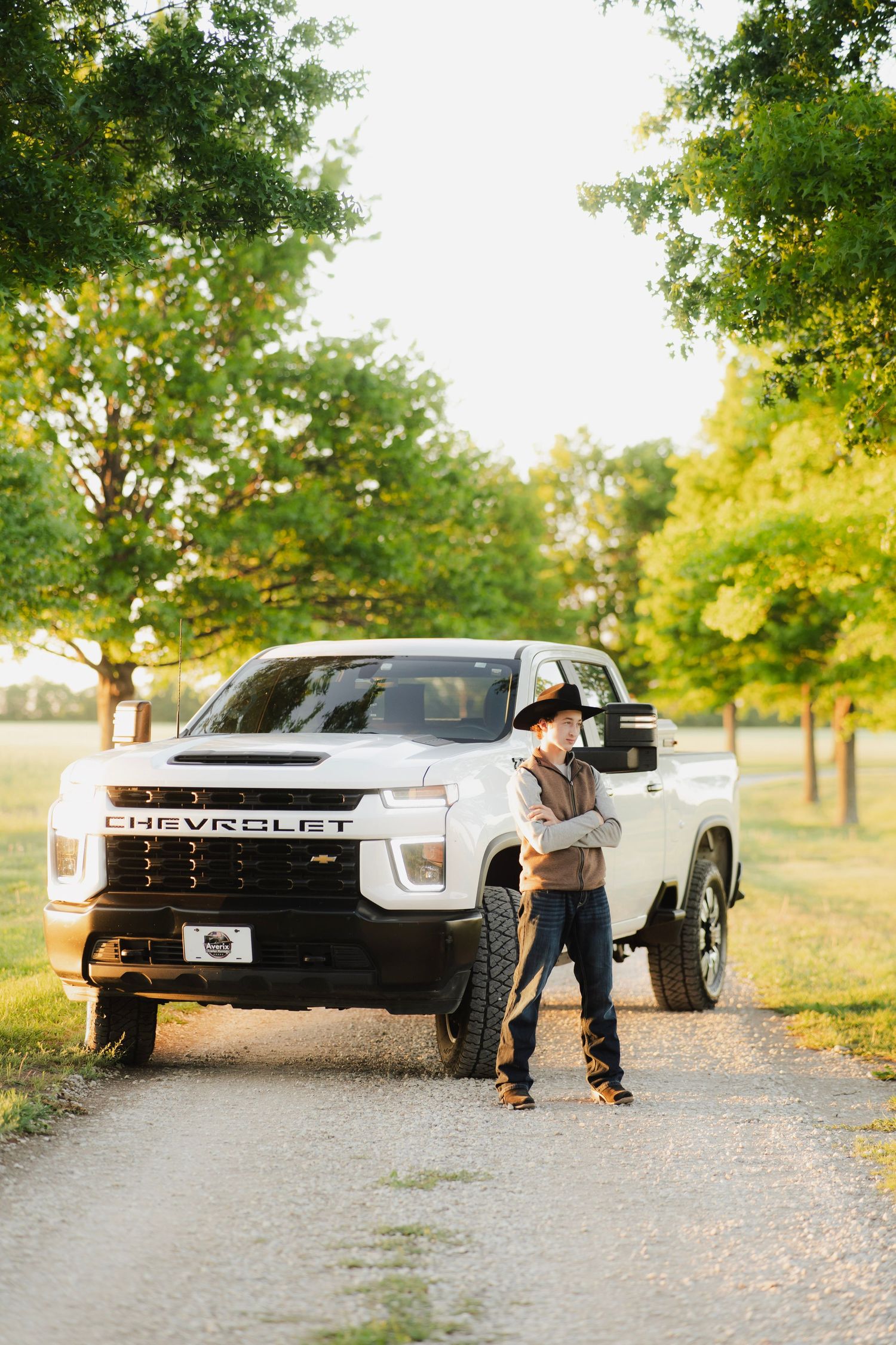 Cowboy in hat leaning against white Chevrolet pickup truck on rural gravel path surrounded by green trees.