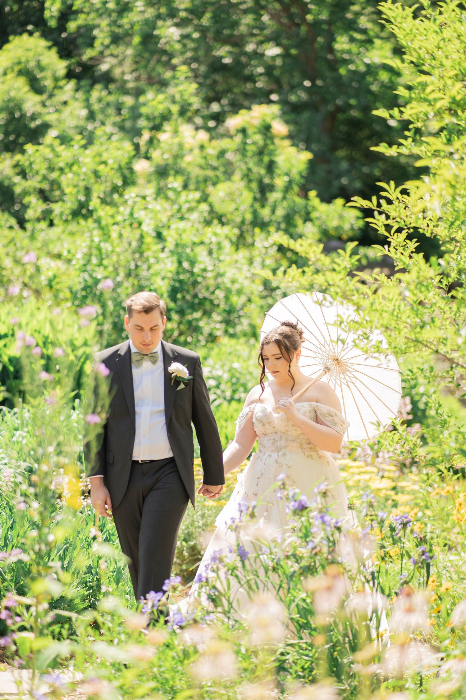 Bride and groom walking hand in hand through lush garden with white parasol on sunny wedding day.