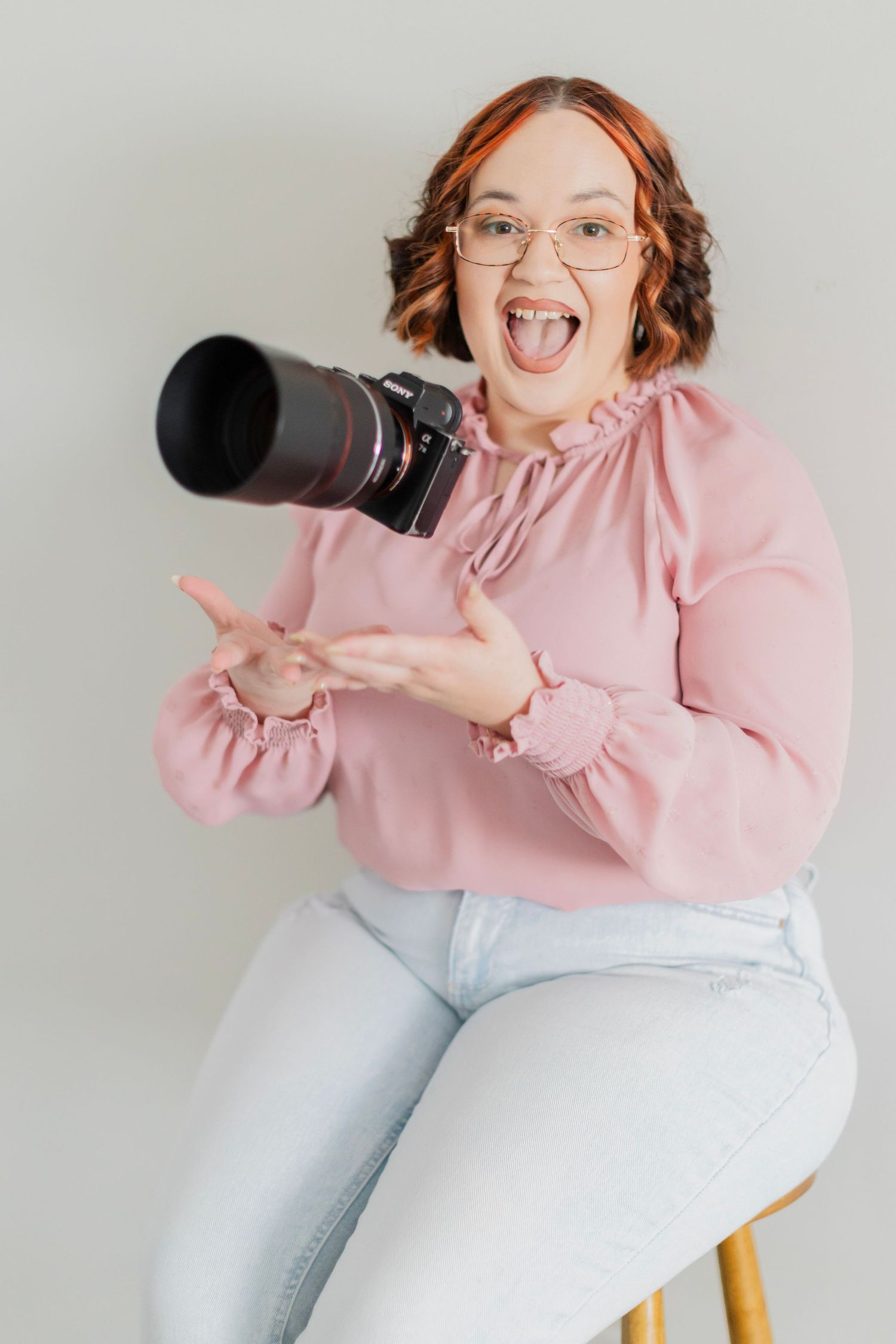 A cheerful woman in pink blouse holds a DSLR camera while sitting on a stool, sticking out her tongue playfully.