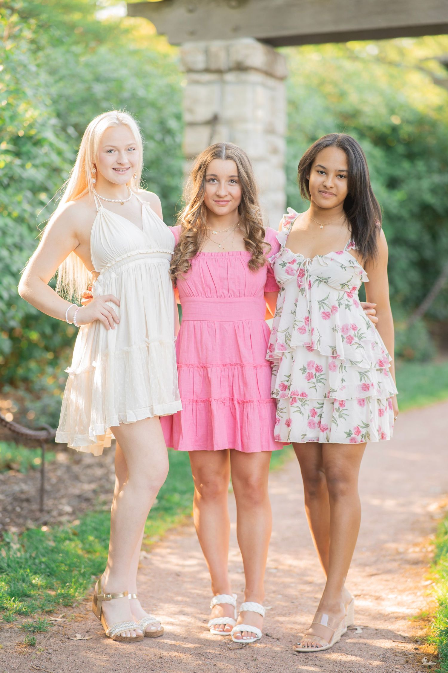 Three teenage girls in colorful summer dresses pose together on a shaded garden path outdoors.
