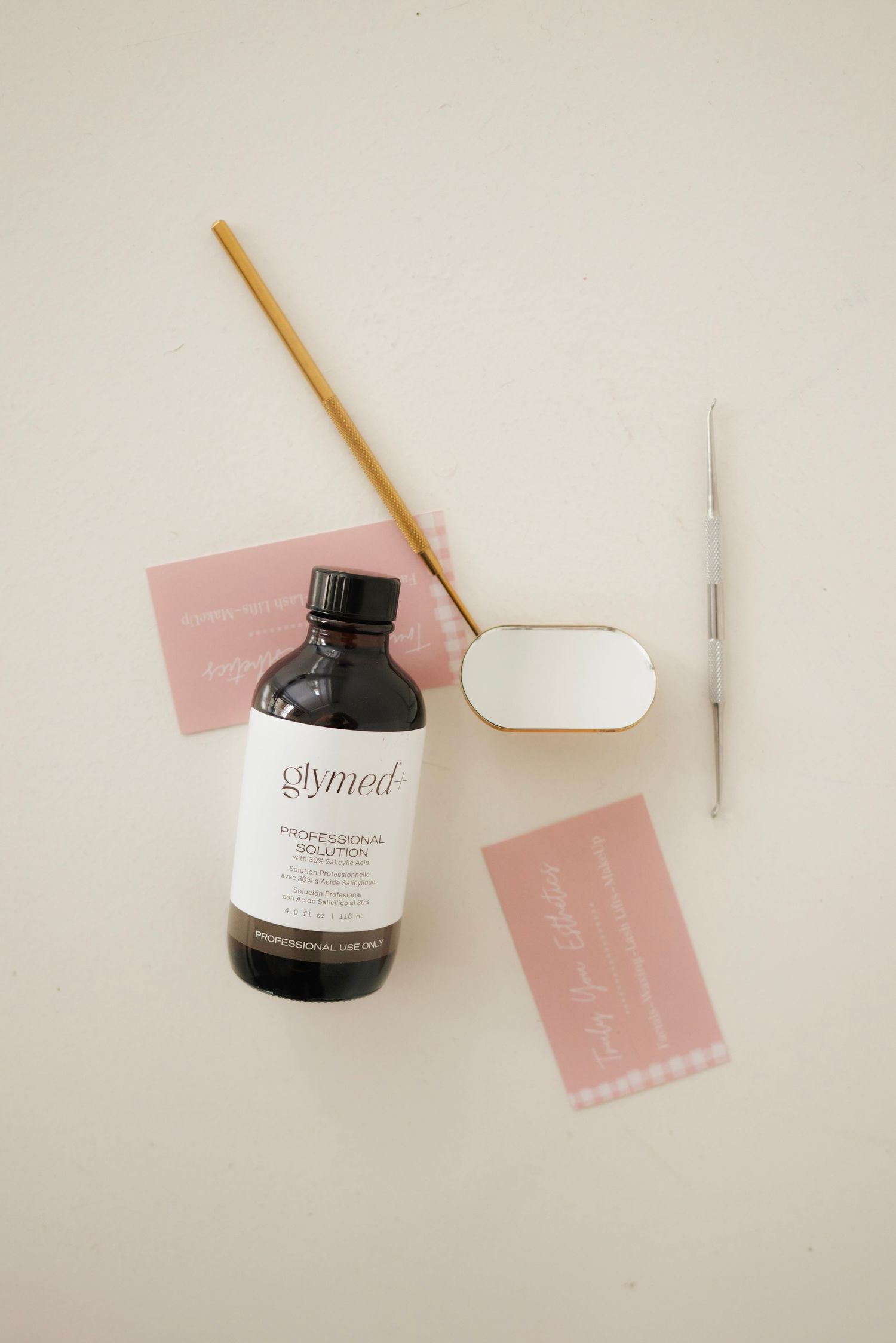 GlyMed Professional Solution bottle with pink business cards, gold brush, mirror, and metal tool on beige background.