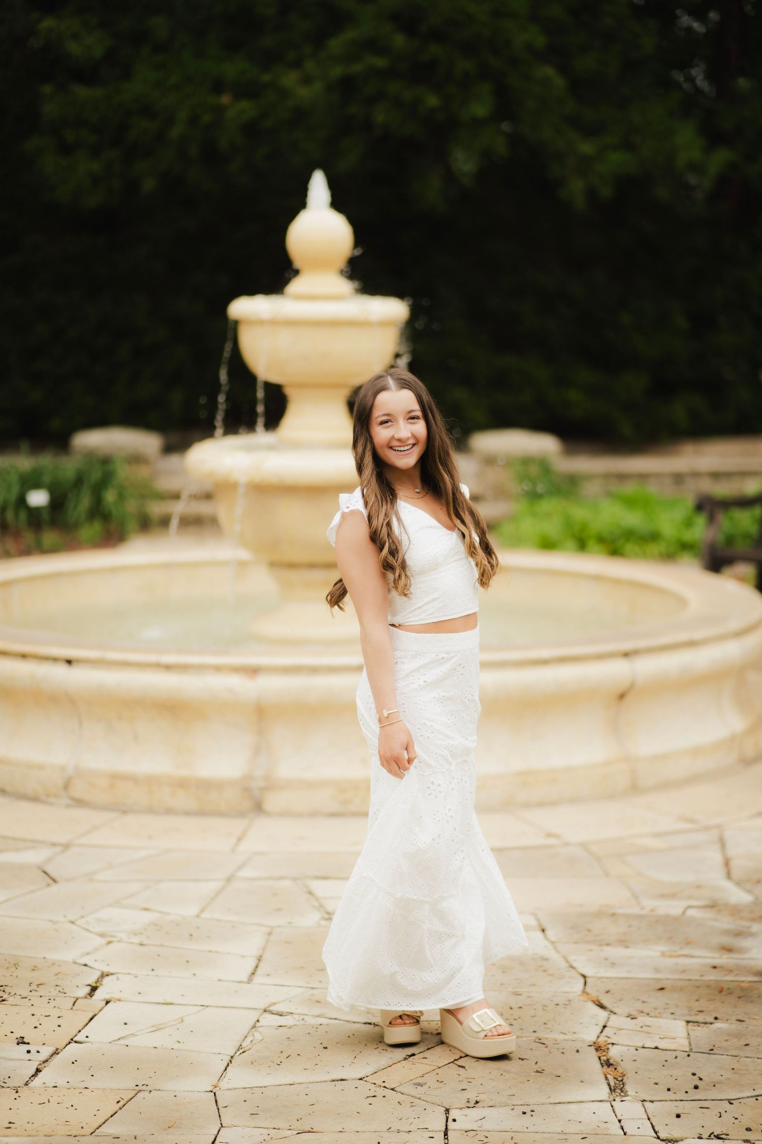 Smiling young woman in white two-piece outfit standing before a stone garden fountain.