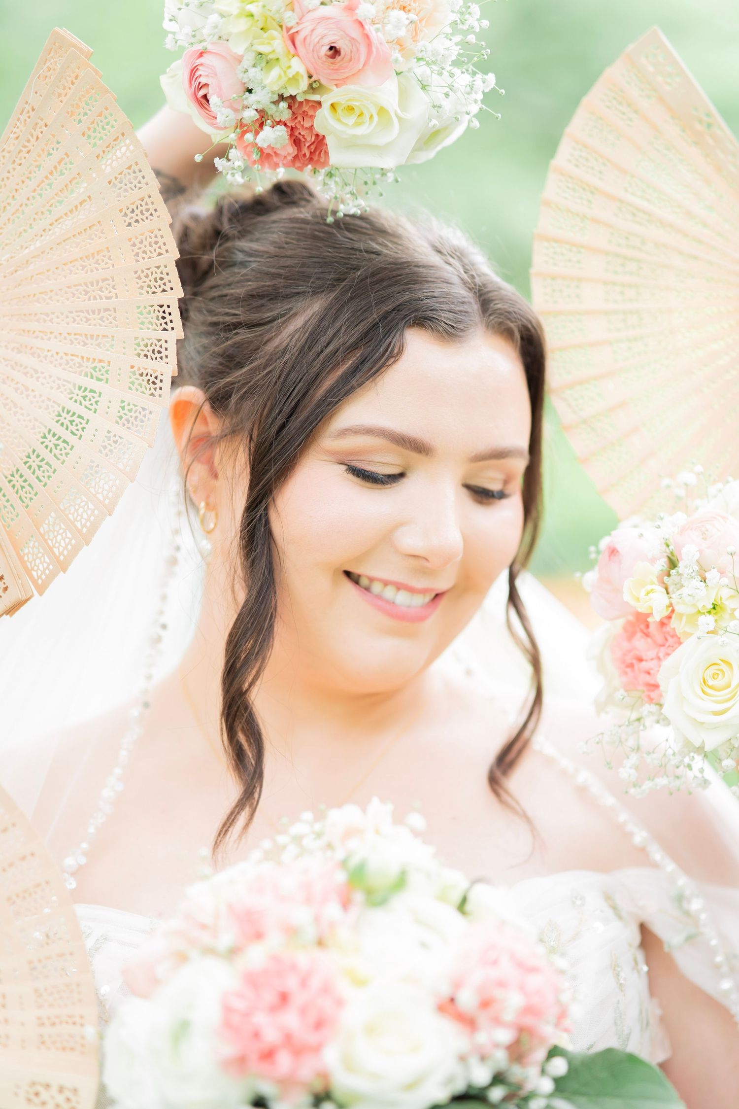 Smiling bride with dark hair holds pink and white floral bouquet, framed by decorative wooden fans.