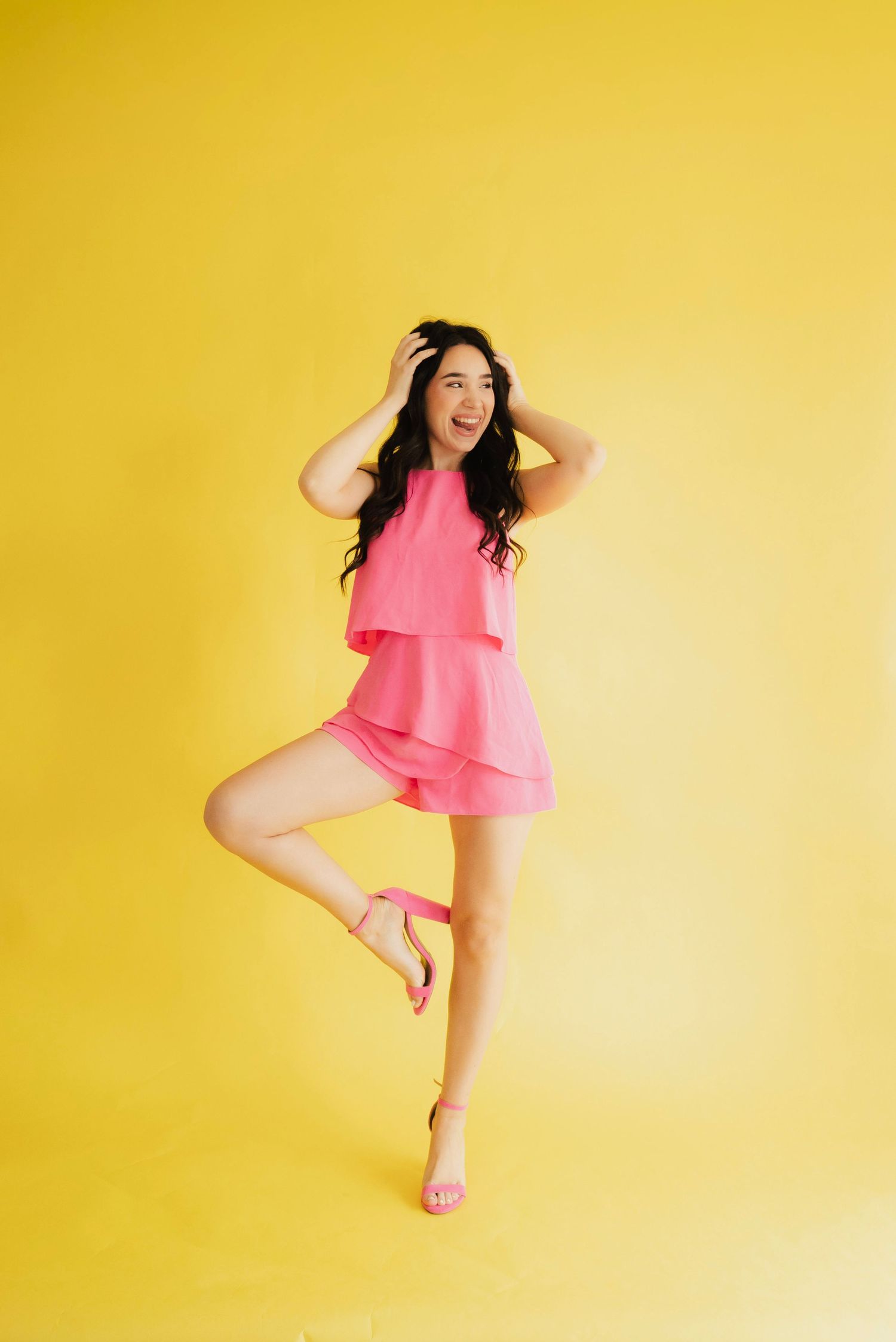 Woman in pink romper and heels striking a fun pose on one leg against a bright yellow background.