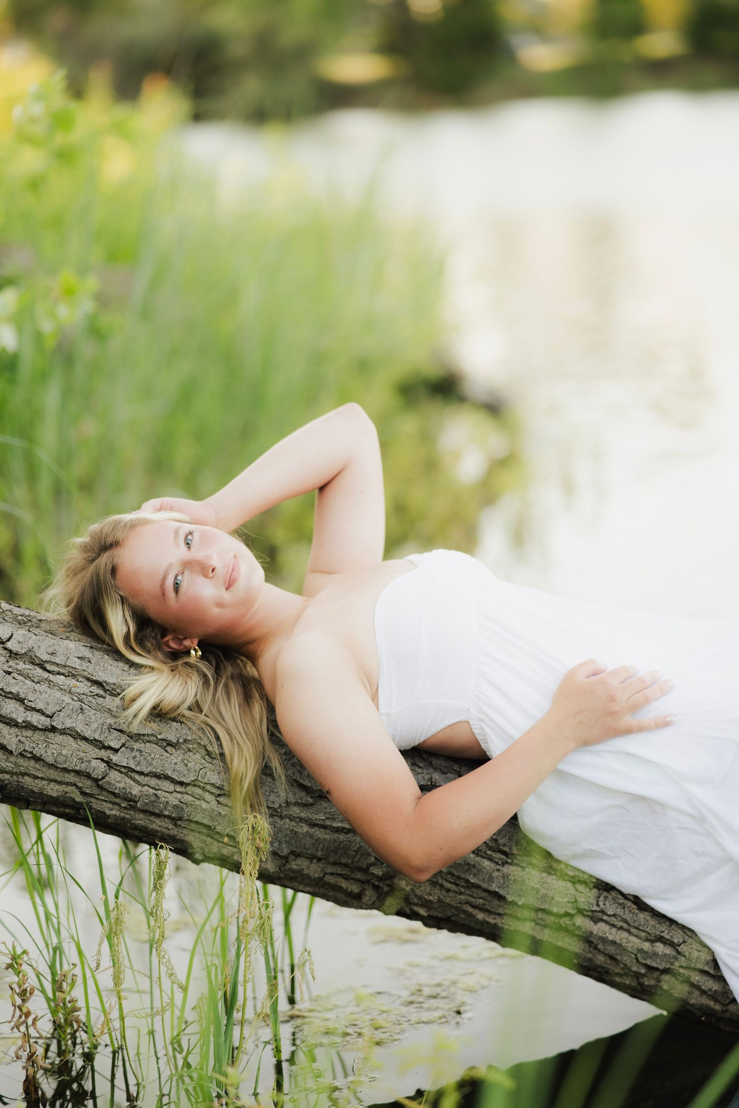Blonde woman in white dress reclining on a log near a serene pond surrounded by lush green grass.