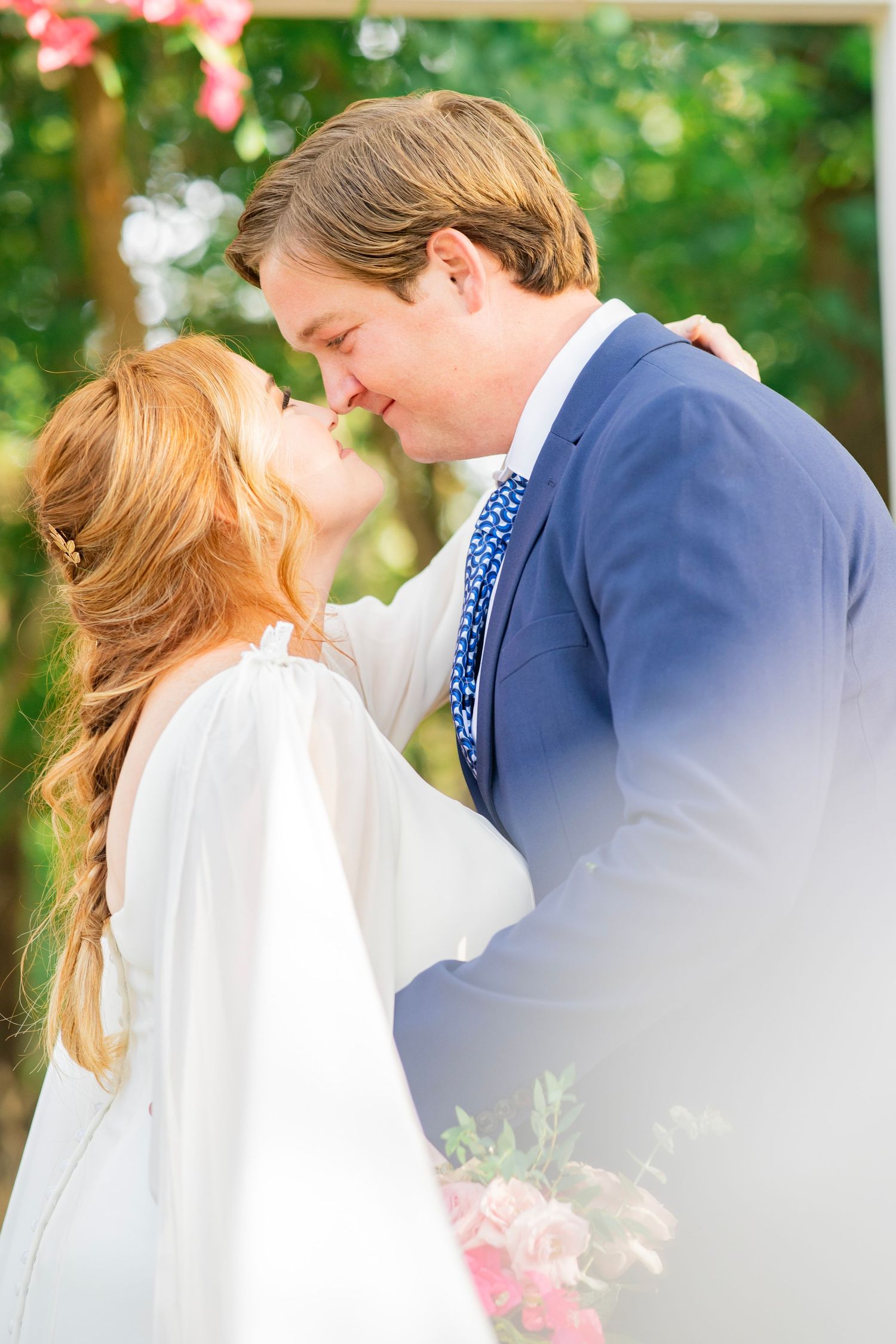 Bride and groom share first kiss outdoors under floral arch at wedding ceremony.