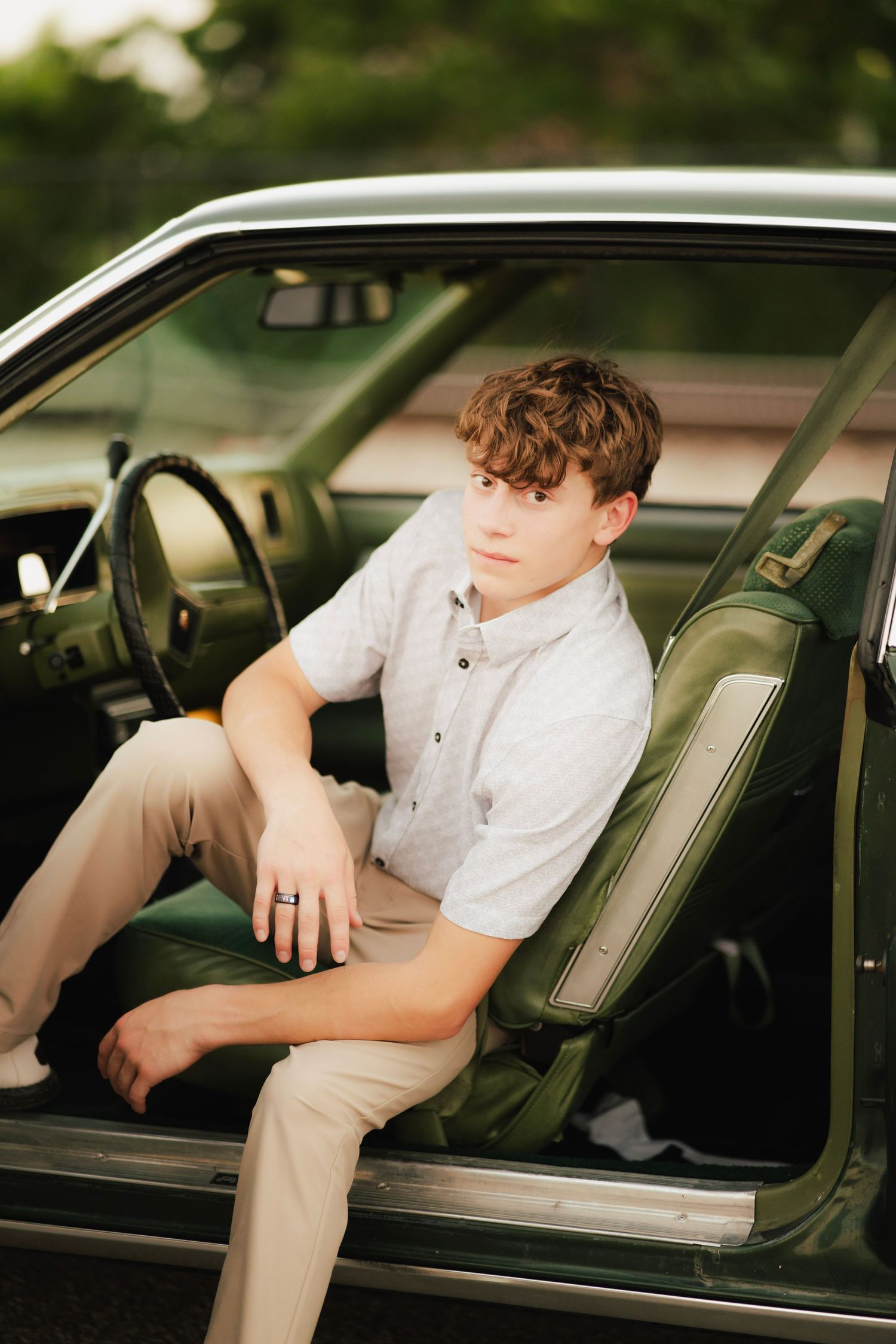 Teen boy in white shirt and khakis sits in vintage green car door for senior portrait photo.