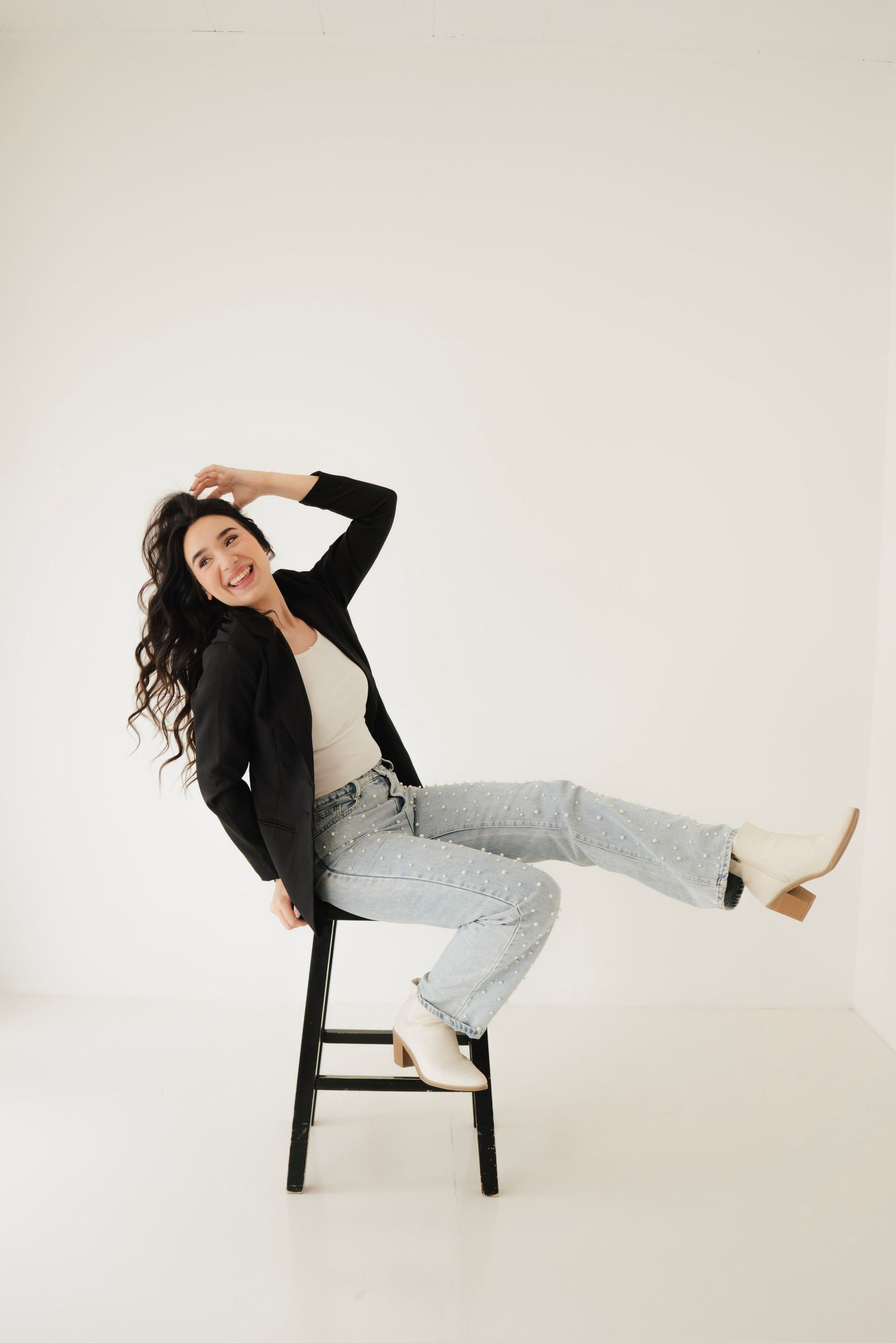 Smiling woman with curly hair sits on black chair, wearing black blazer, light jeans, and beige heels against white background.