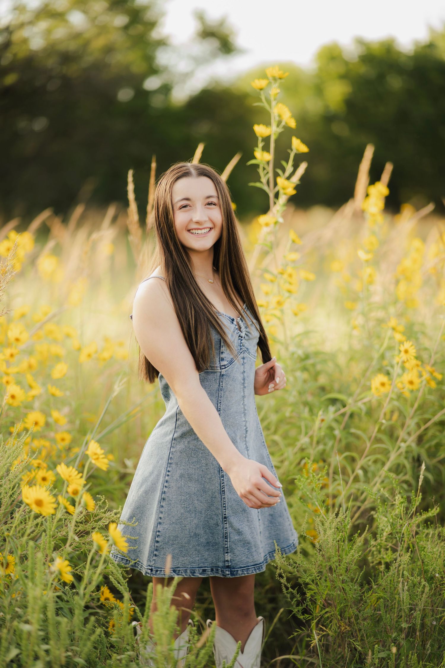 Smiling brunette teen in denim dress and white boots stands among tall yellow wildflowers in golden sunlight.