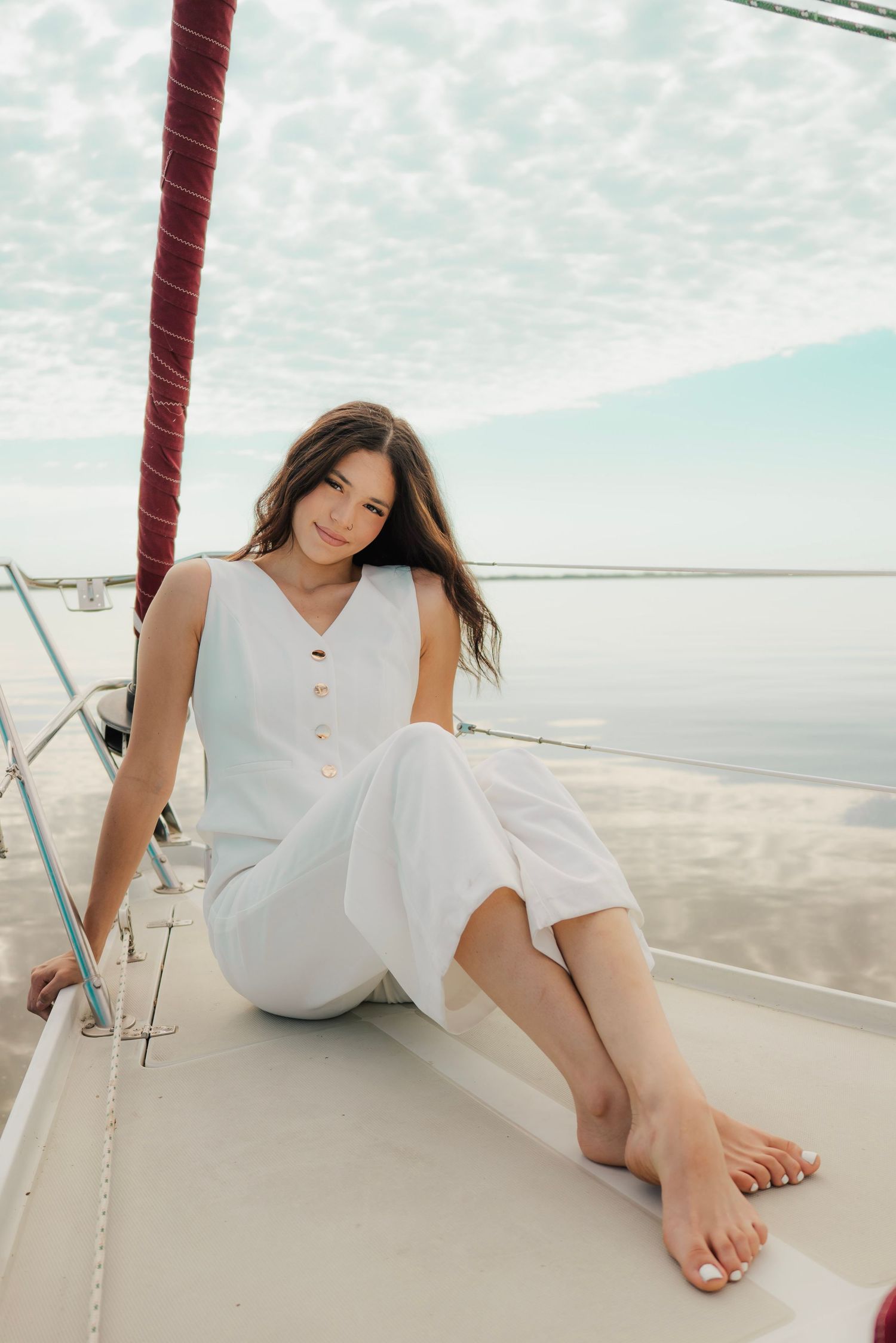 Smiling young woman in white linen outfit sits barefoot on sailboat deck with ocean and cloudy sky behind her.