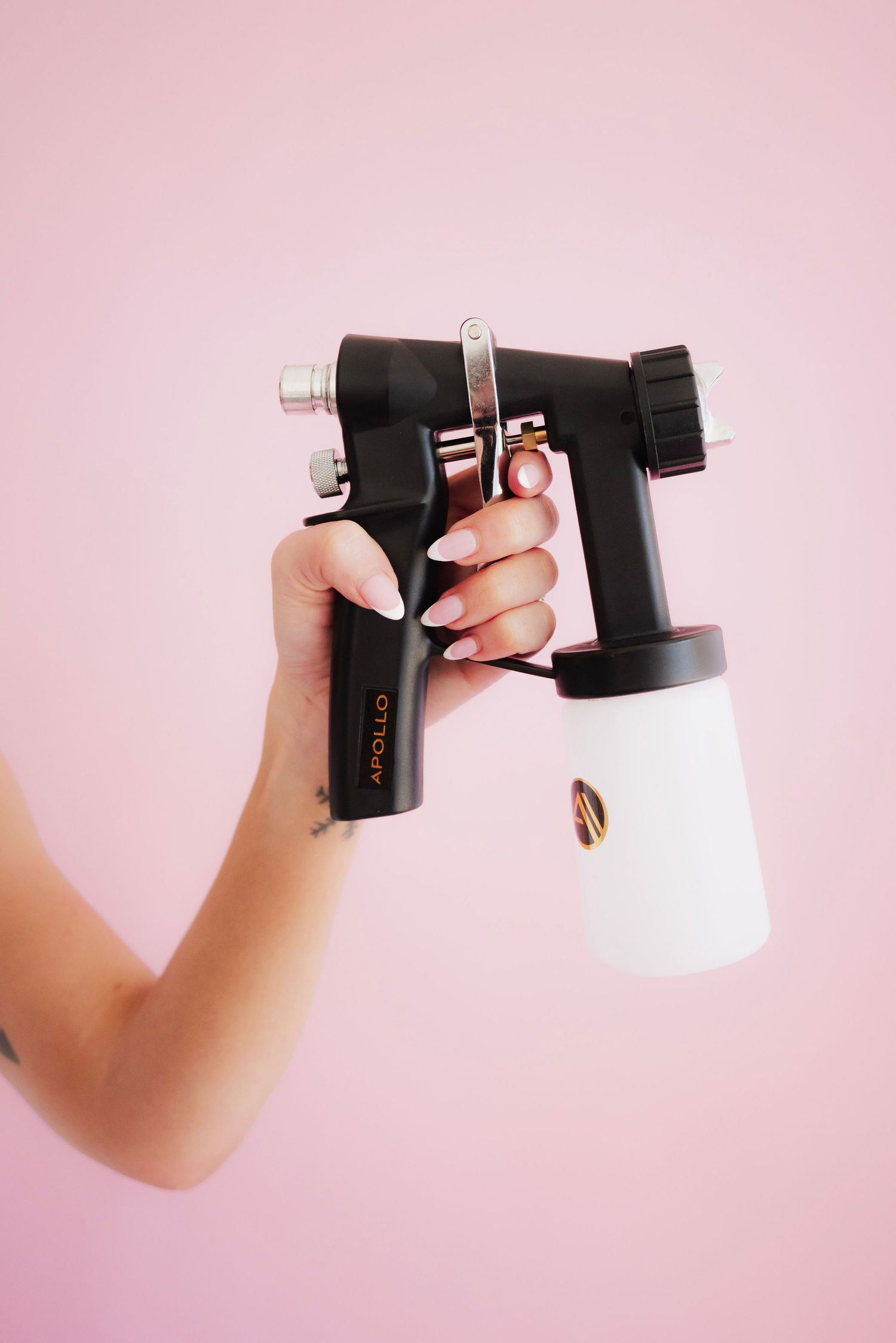 Hand holding an Apollo spray tan gun with white cup against a pink background.