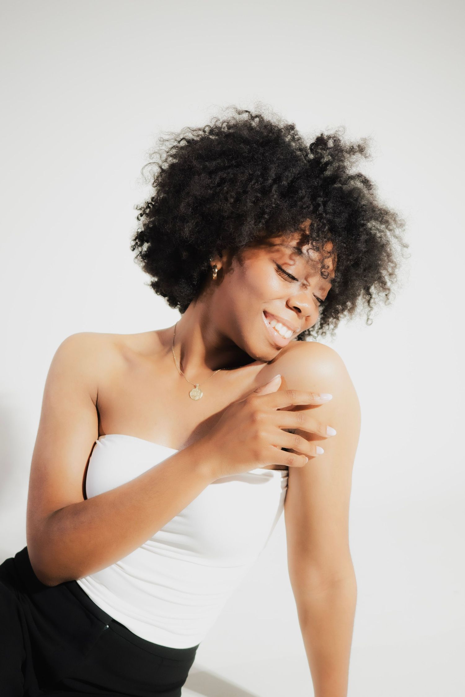 Smiling Black woman with natural afro hair wearing a white tube top and gold necklace, touching her shoulder against white background.