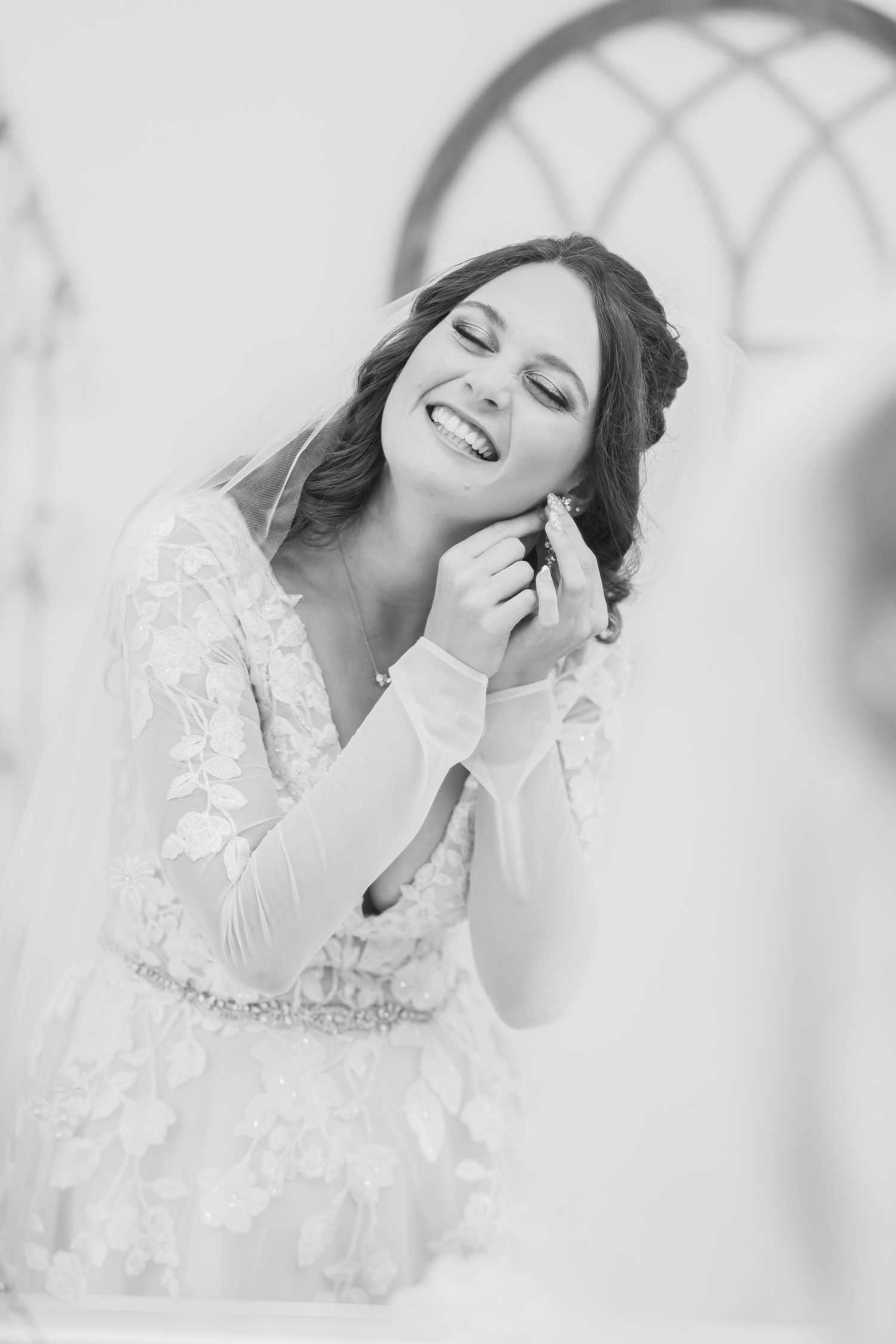 Black and white portrait of smiling bride in lace gown putting on earrings.