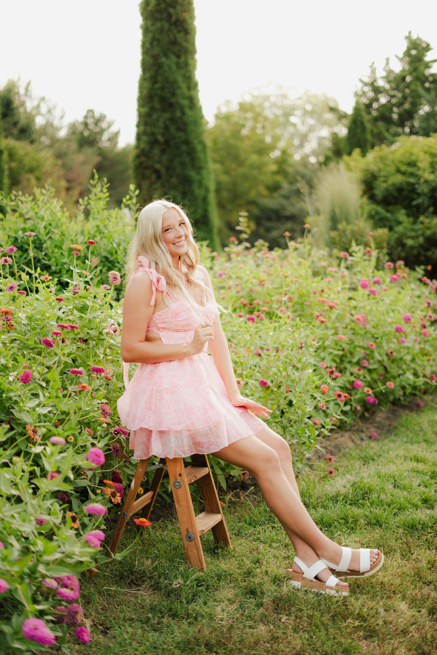 Blonde woman in pink floral dress sitting on wooden stool surrounded by colorful garden flowers.