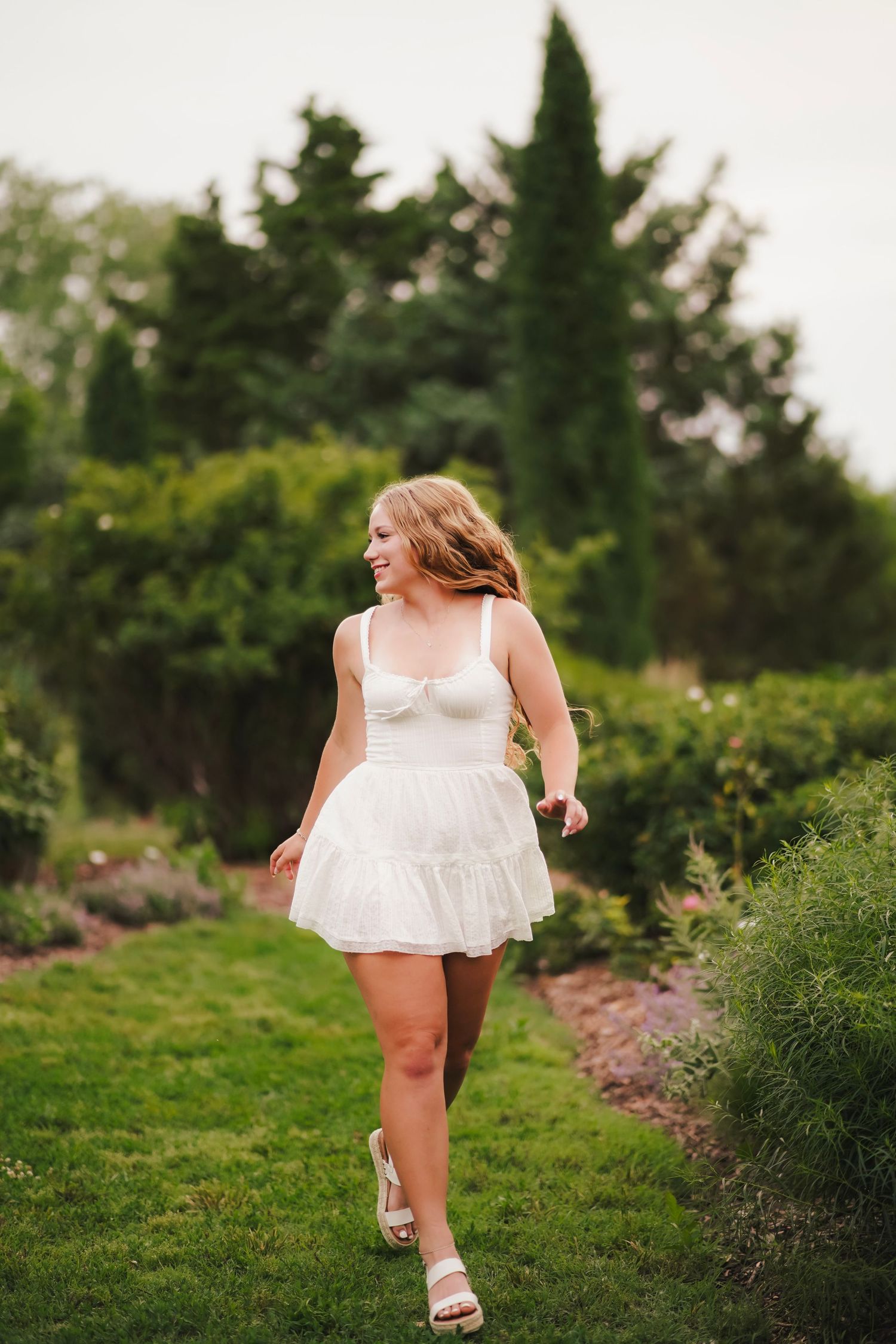 Smiling blonde teen in white mini dress walking through lush green garden path.