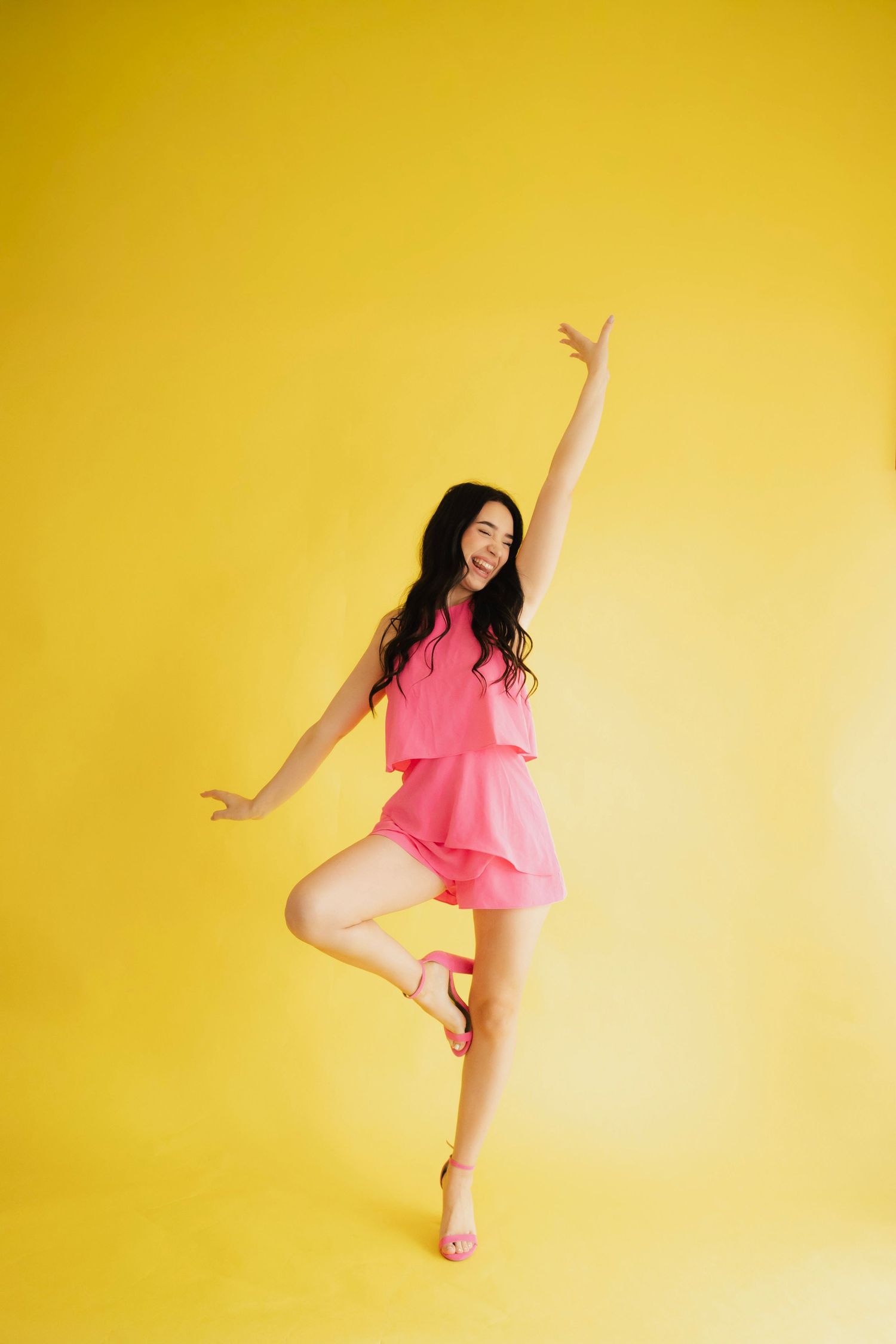 A joyful young woman in a pink outfit balances on one leg with arms raised against a bright yellow background.