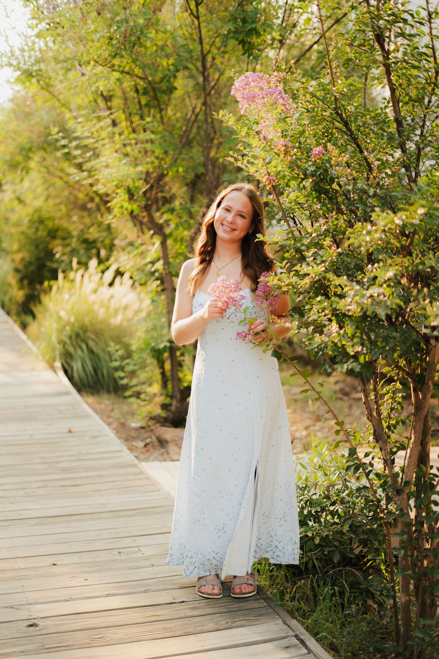 Young woman in white maxi dress holding pink flowers on a wooden boardwalk path surrounded by lush greenery.