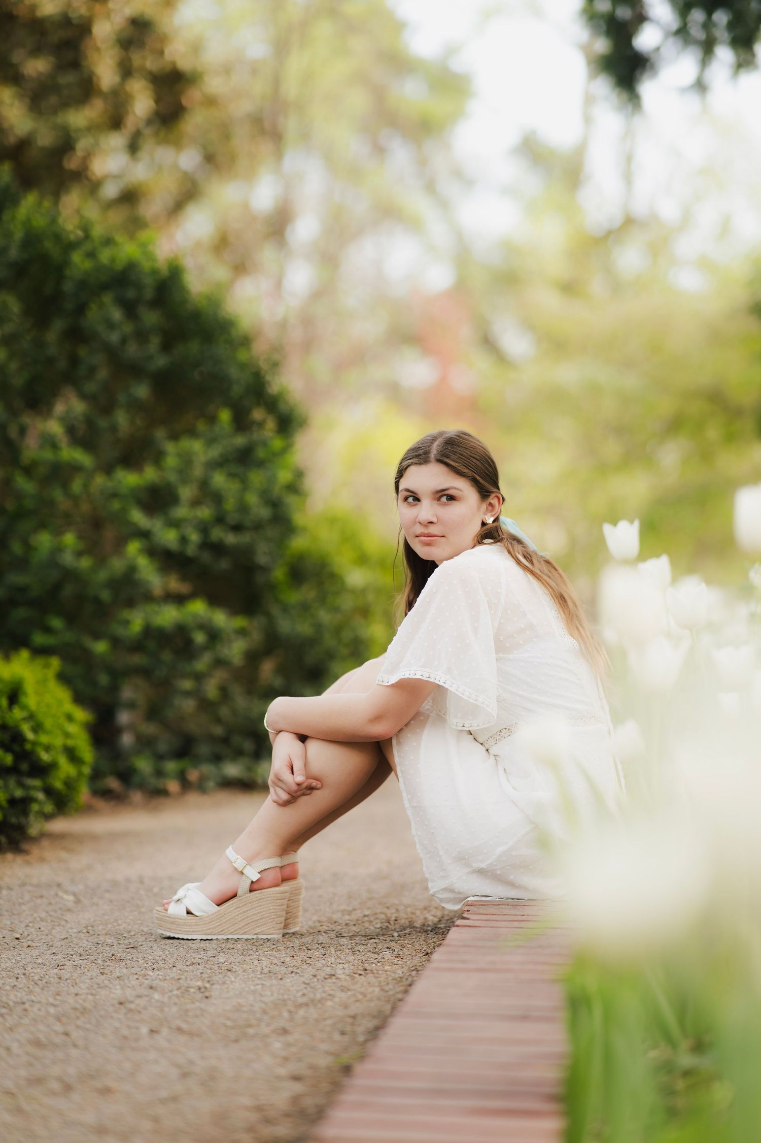 Young brunette woman in white dress crouches beside white tulips on garden path in spring park.