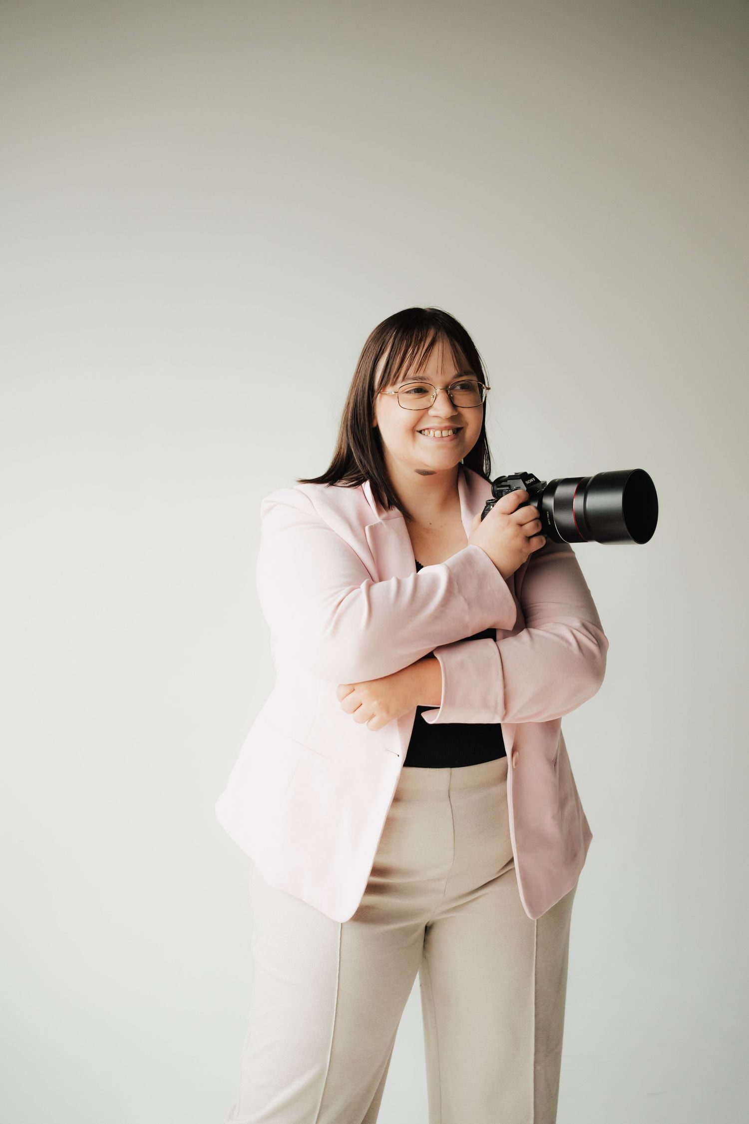 Smiling female photographer in pink blazer holds DSLR camera against light gray background.