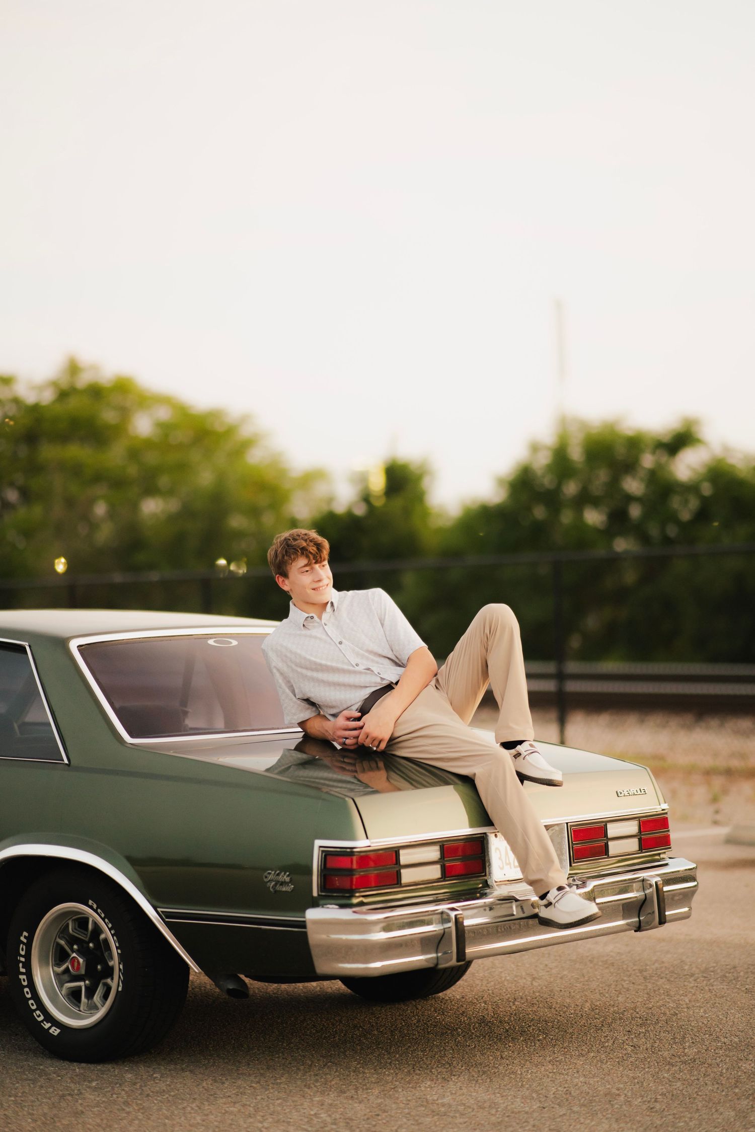 Young man in khakis reclines on trunk of vintage green classic car at golden hour.
