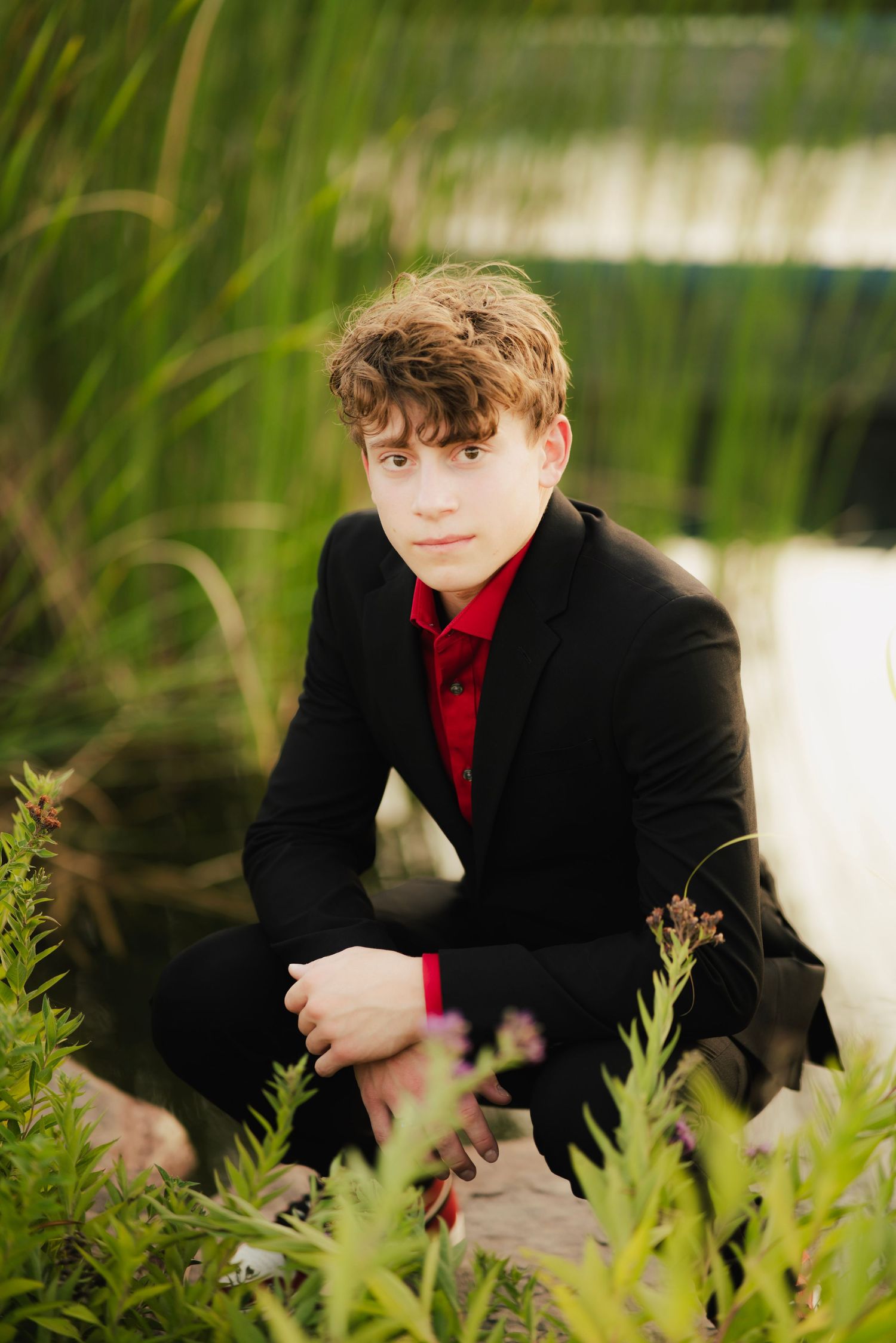 Young man in black suit and red shirt sitting among green plants near a pond in an outdoor portrait.