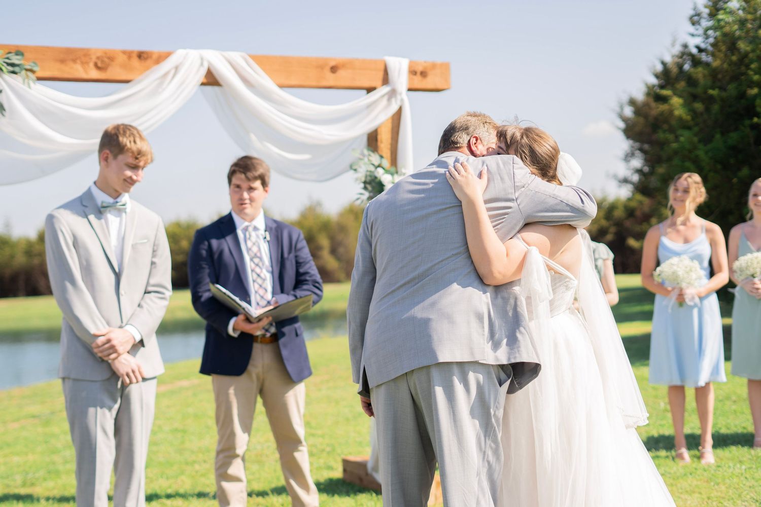Bride and groom embrace at outdoor wedding ceremony under wooden arch with white draping.