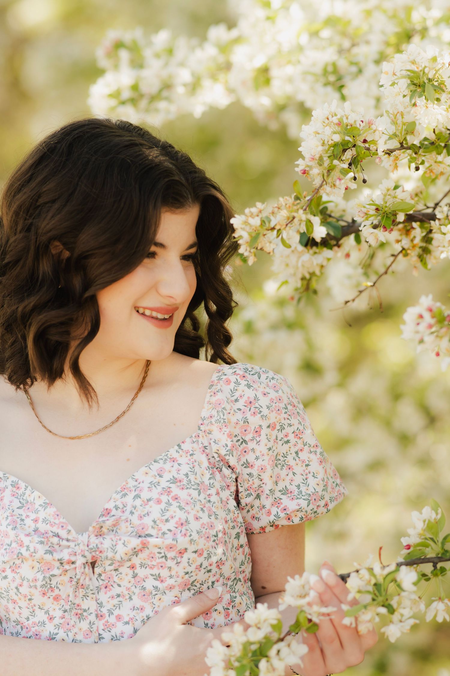 Brunette teen girl in floral dress smiling beside blooming white apple tree in sunlight.