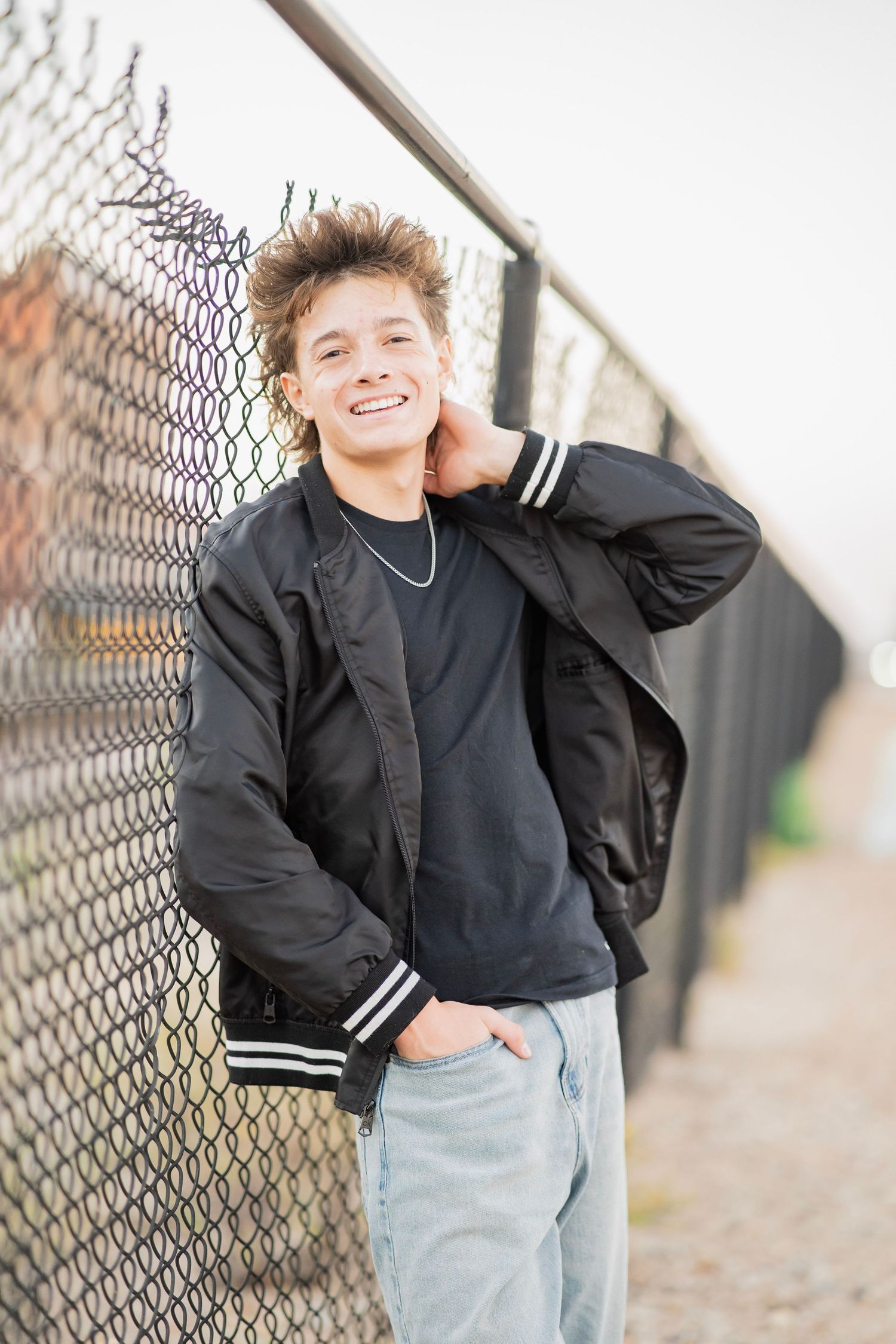 Smiling teenage boy in black bomber jacket and jeans leaning against chain-link fence outdoors.