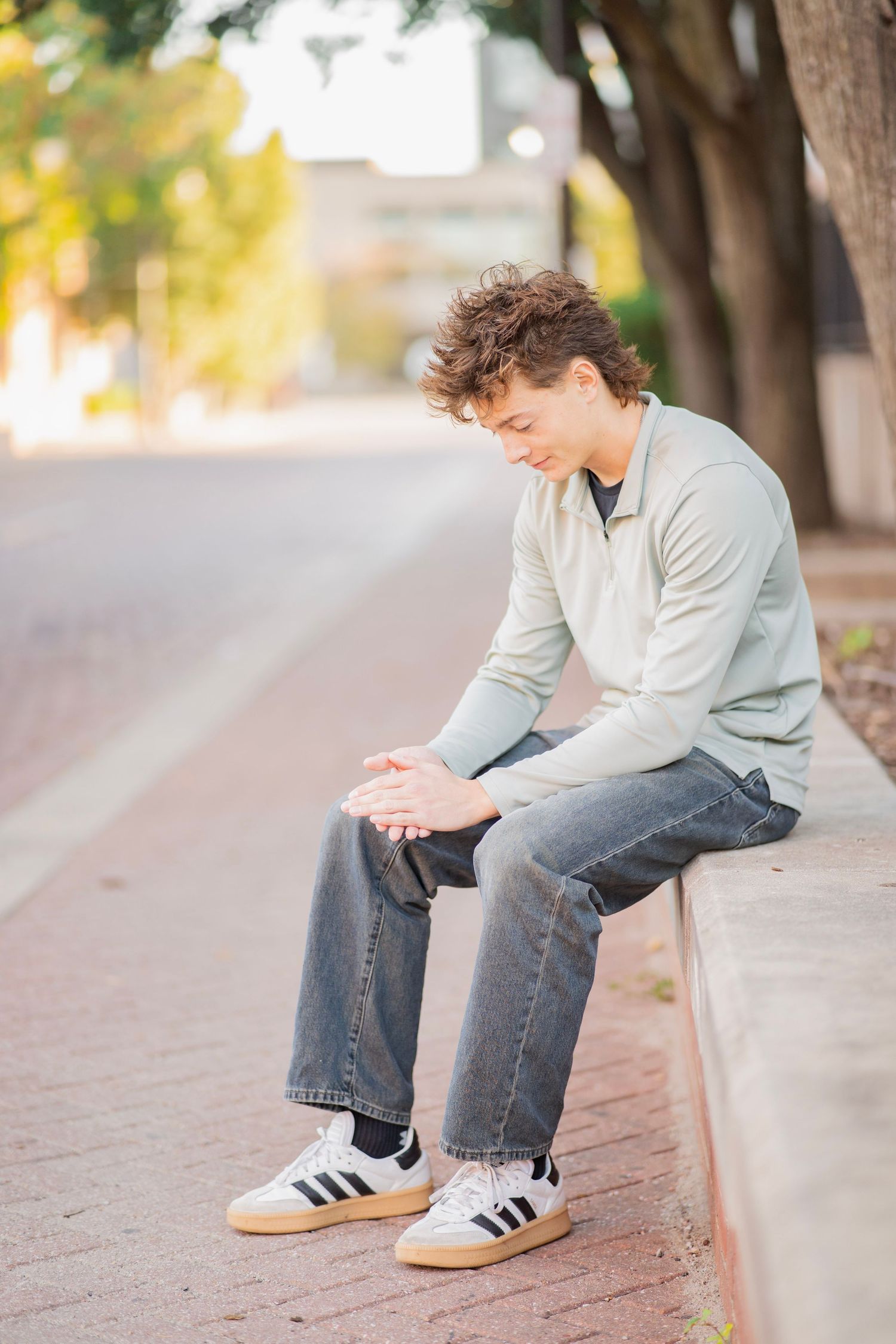 Young man with curly hair sitting on a curb in jeans and sneakers, looking down thoughtfully on a tree-lined street.