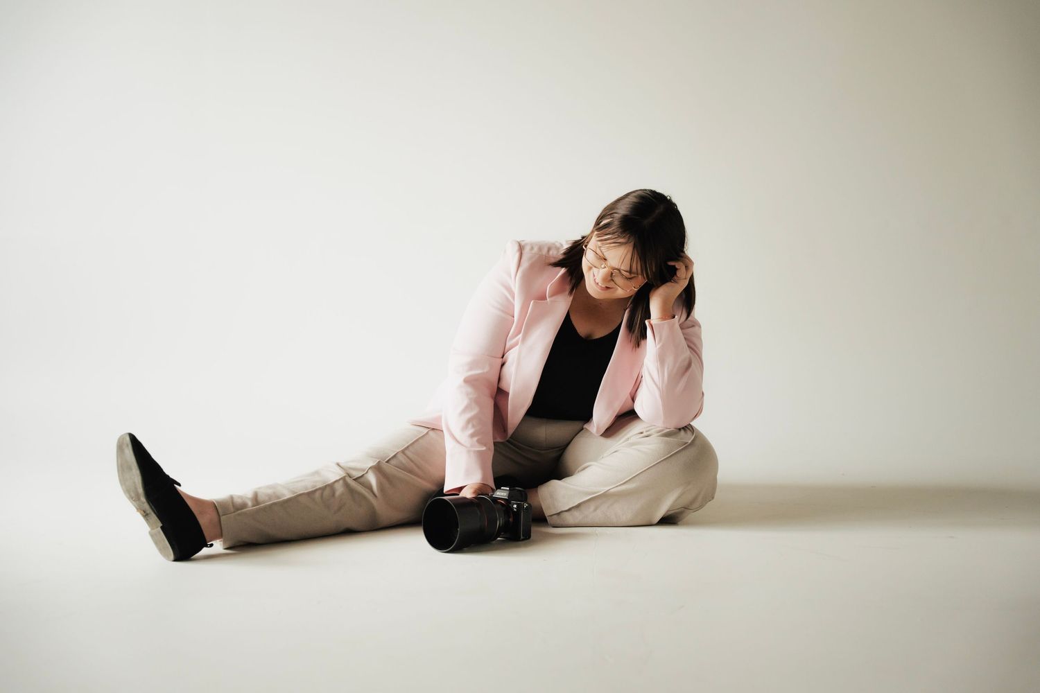 Young woman in pink blazer sits on floor looking down at camera against white background.