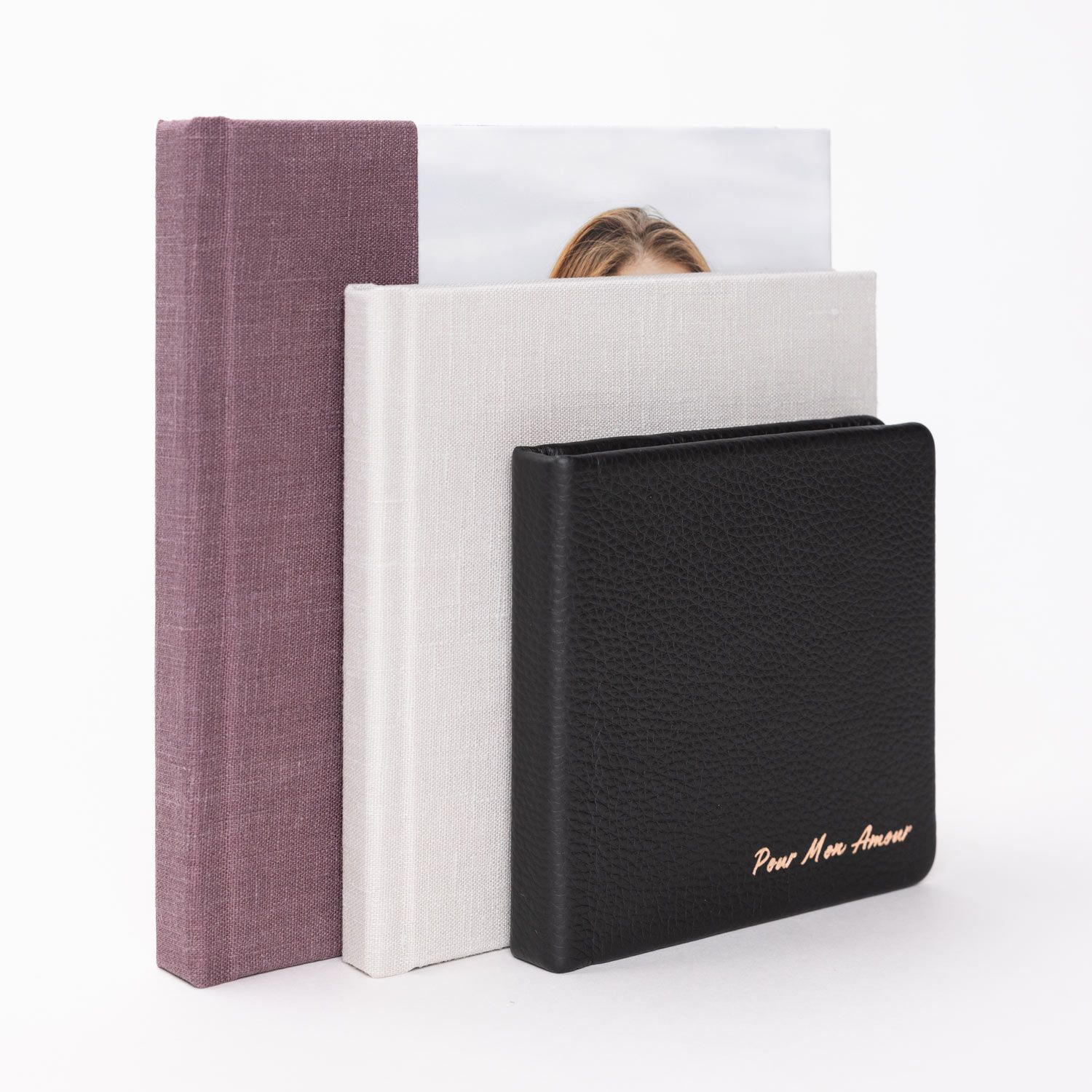 Three photo albums in purple linen, white linen, and black leather with gold foil text standing upright.