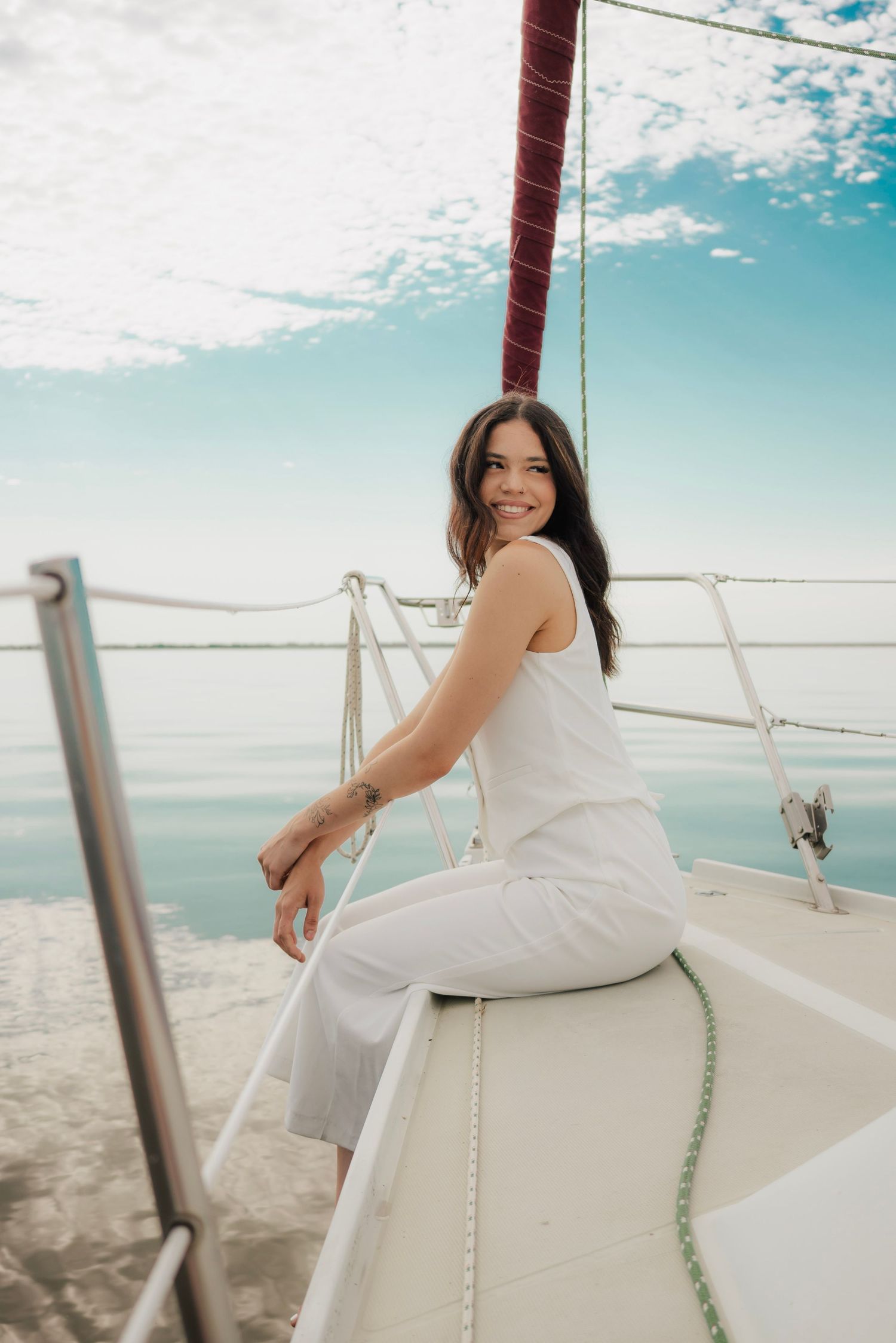 Smiling brunette woman in white outfit sitting on bow of sailboat on calm water.